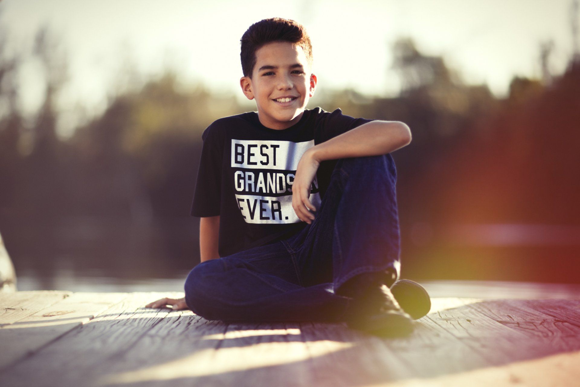 A young boy is sitting on a dock wearing a shirt that says `` best grandpa ever ''.