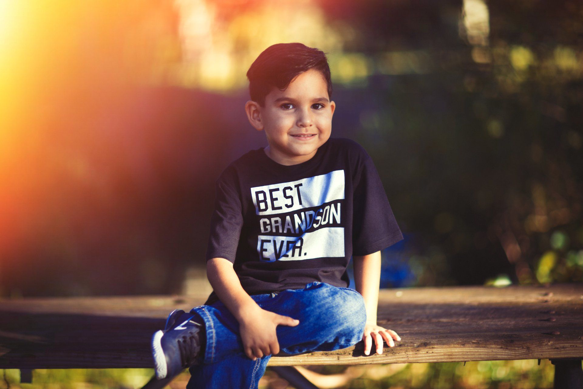 A young boy is sitting on a bench wearing a shirt that says `` best grandson ever ''.