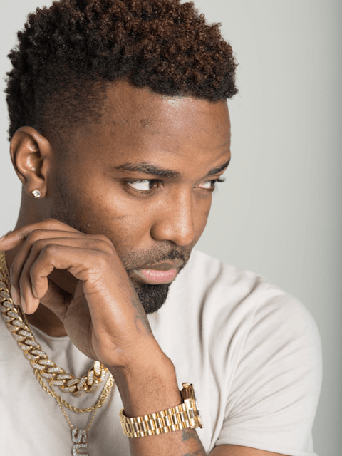 A man wearing a white shirt and a gold chain has his hand on his chin.