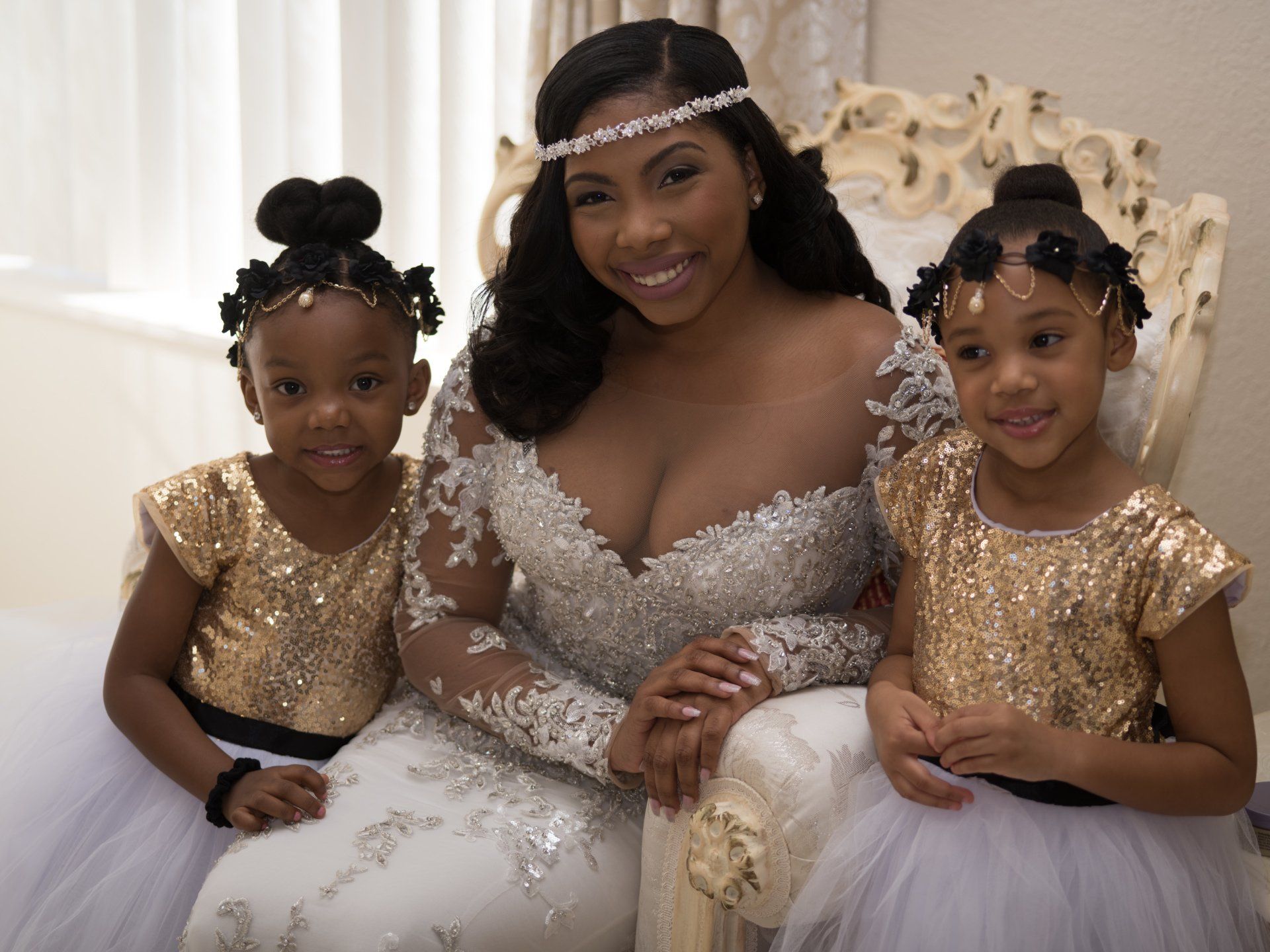 A woman in a wedding dress is sitting on a chair with two flower girls.