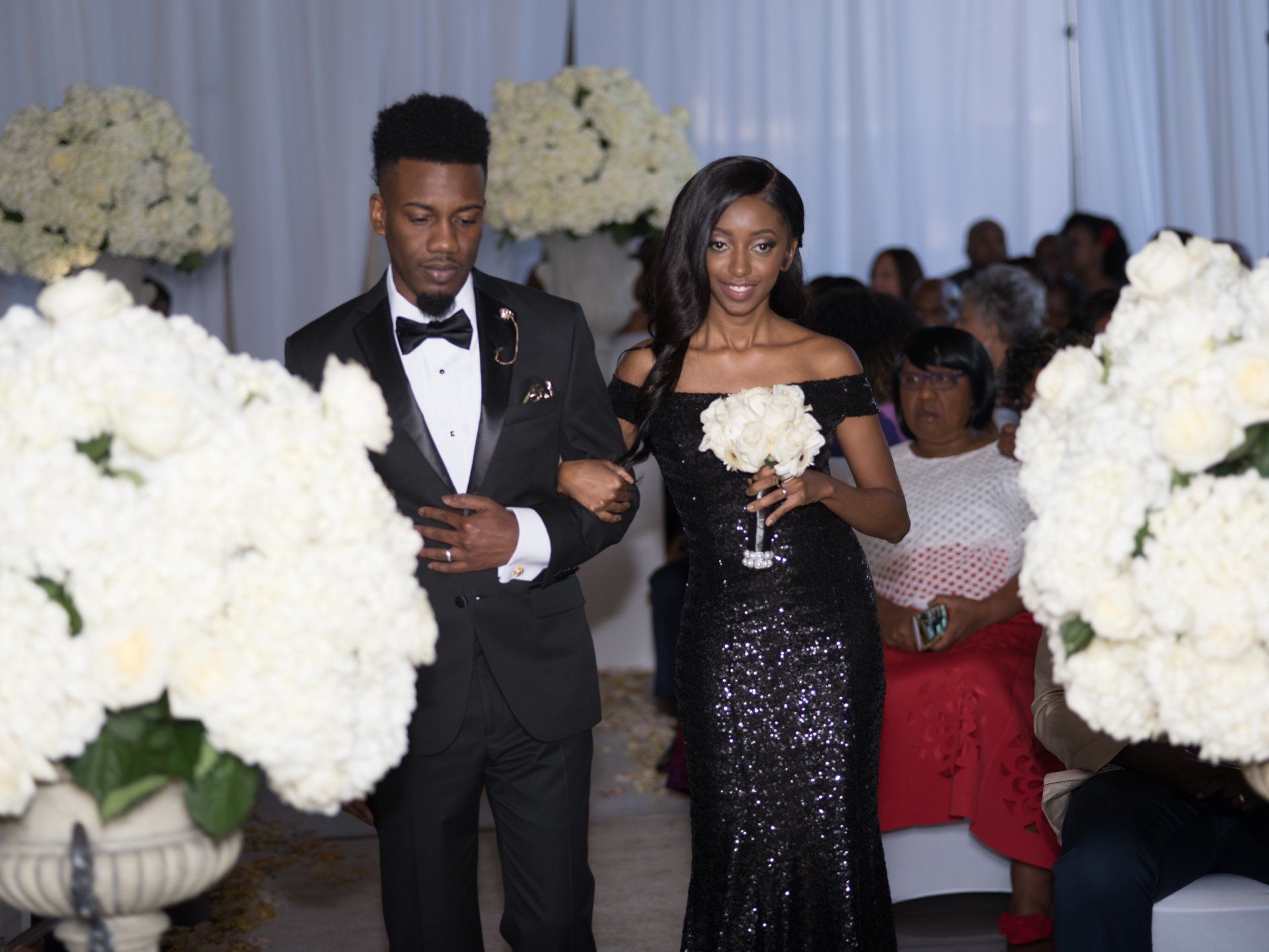 A man in a tuxedo is walking down the aisle with a woman in a black dress.