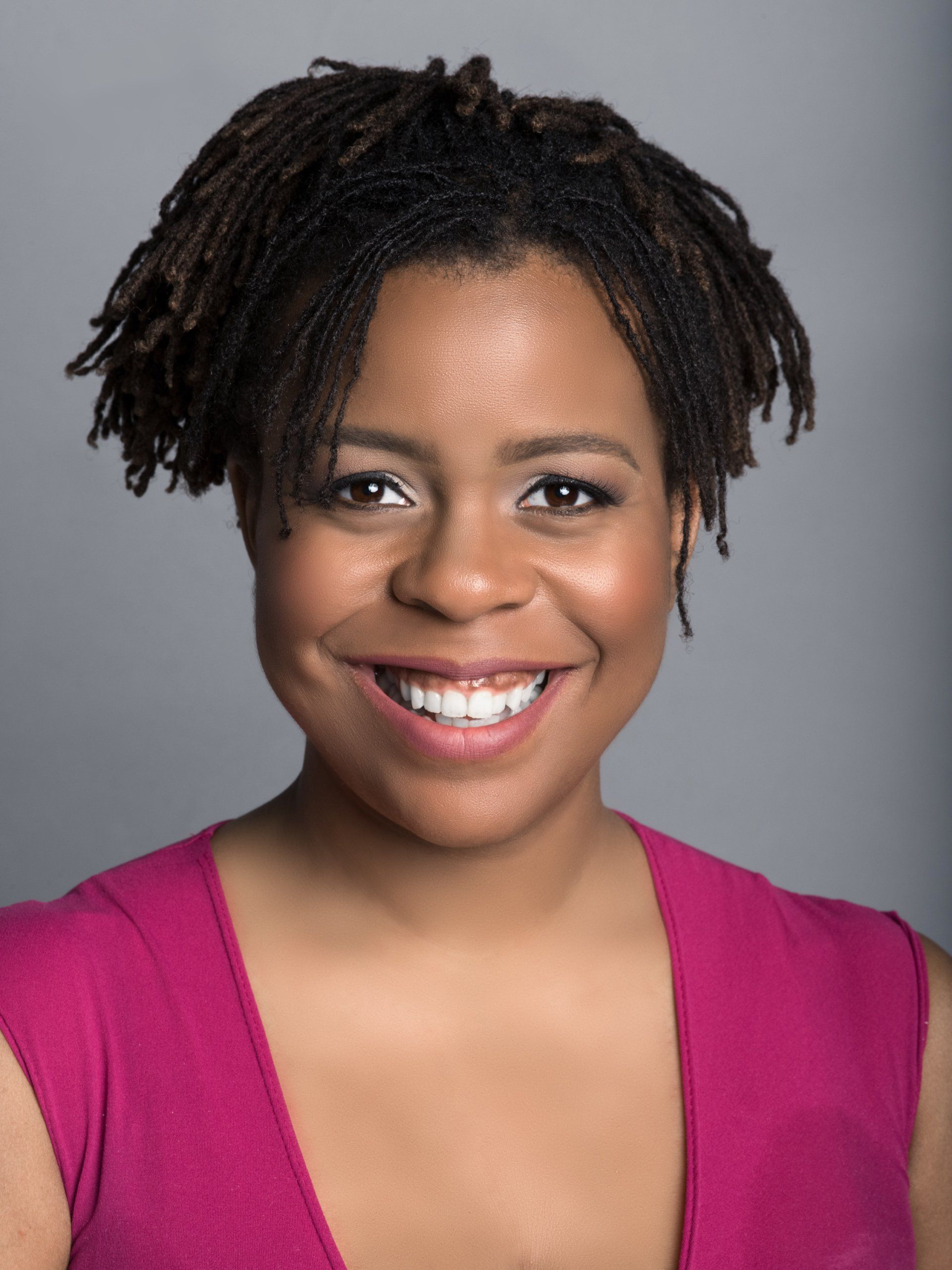 A woman with dreadlocks is smiling and wearing a pink shirt