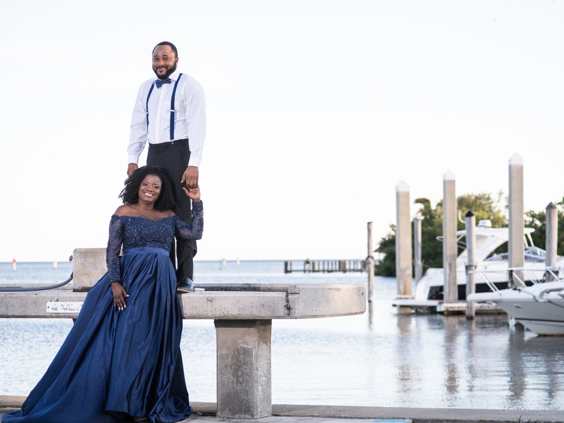 A pregnant woman in a blue dress is sitting on a dock next to a man.