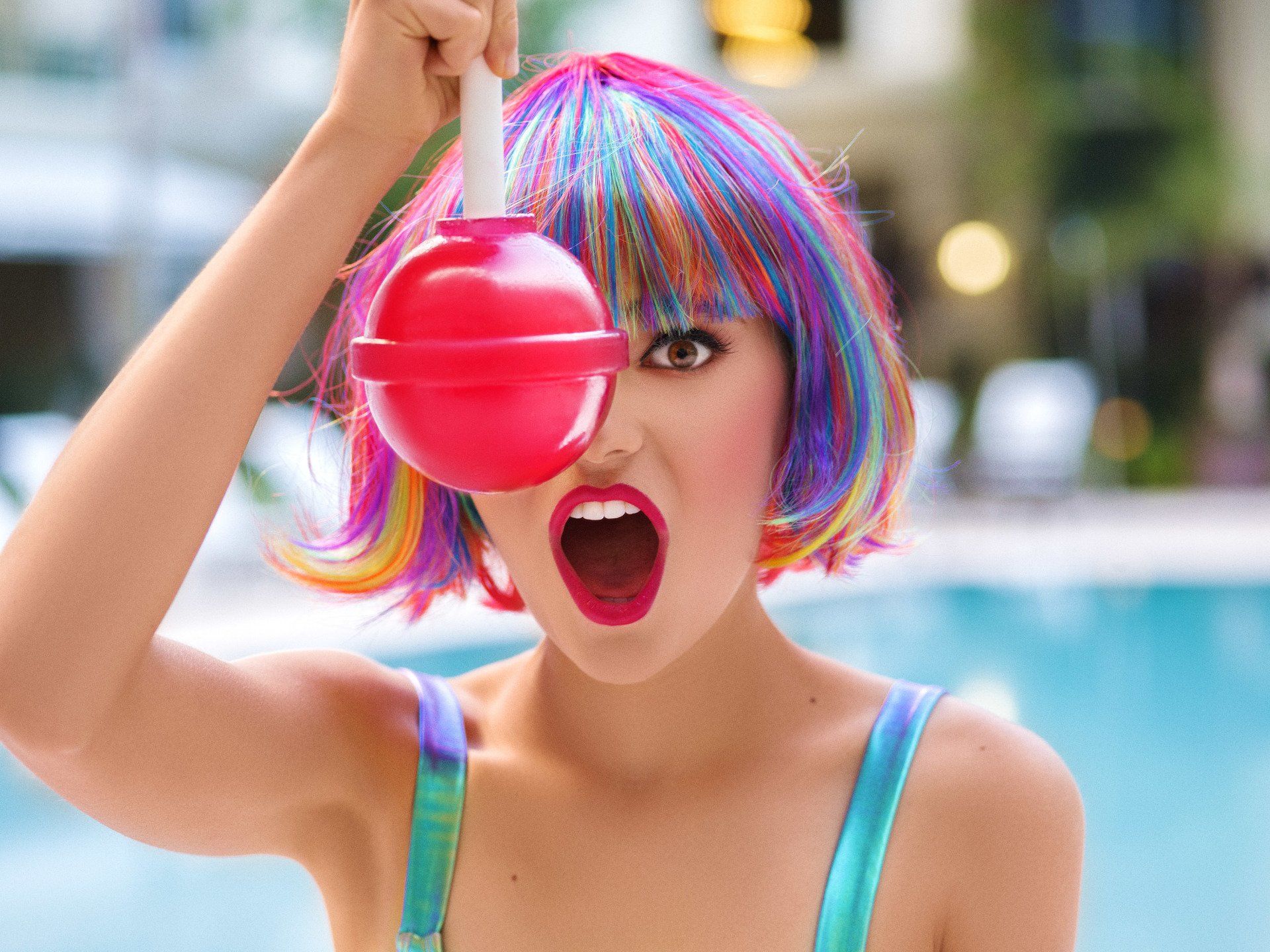 A woman in a colorful wig is holding a lollipop over her eye
