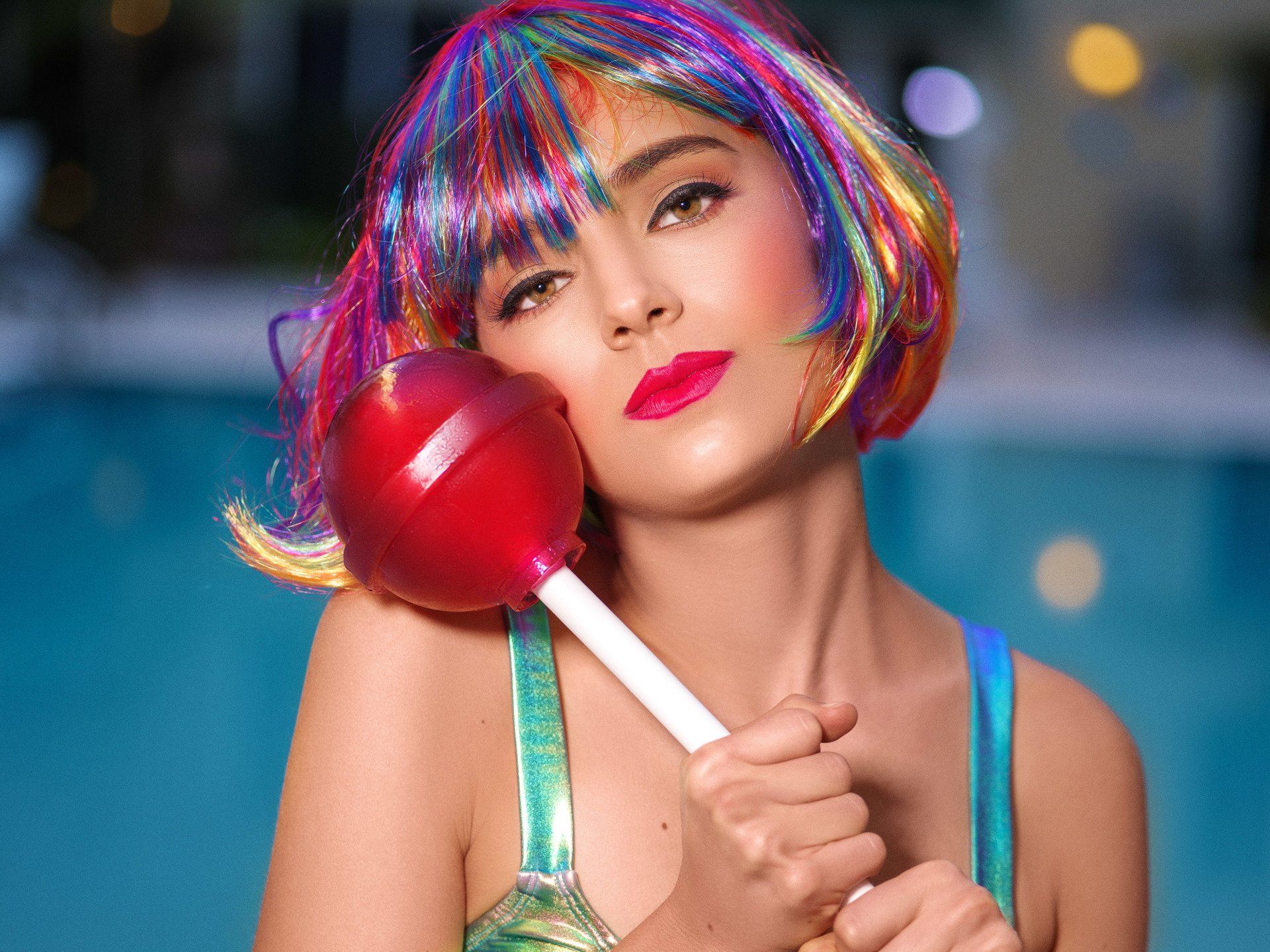A woman in a colorful wig is holding a red lollipop