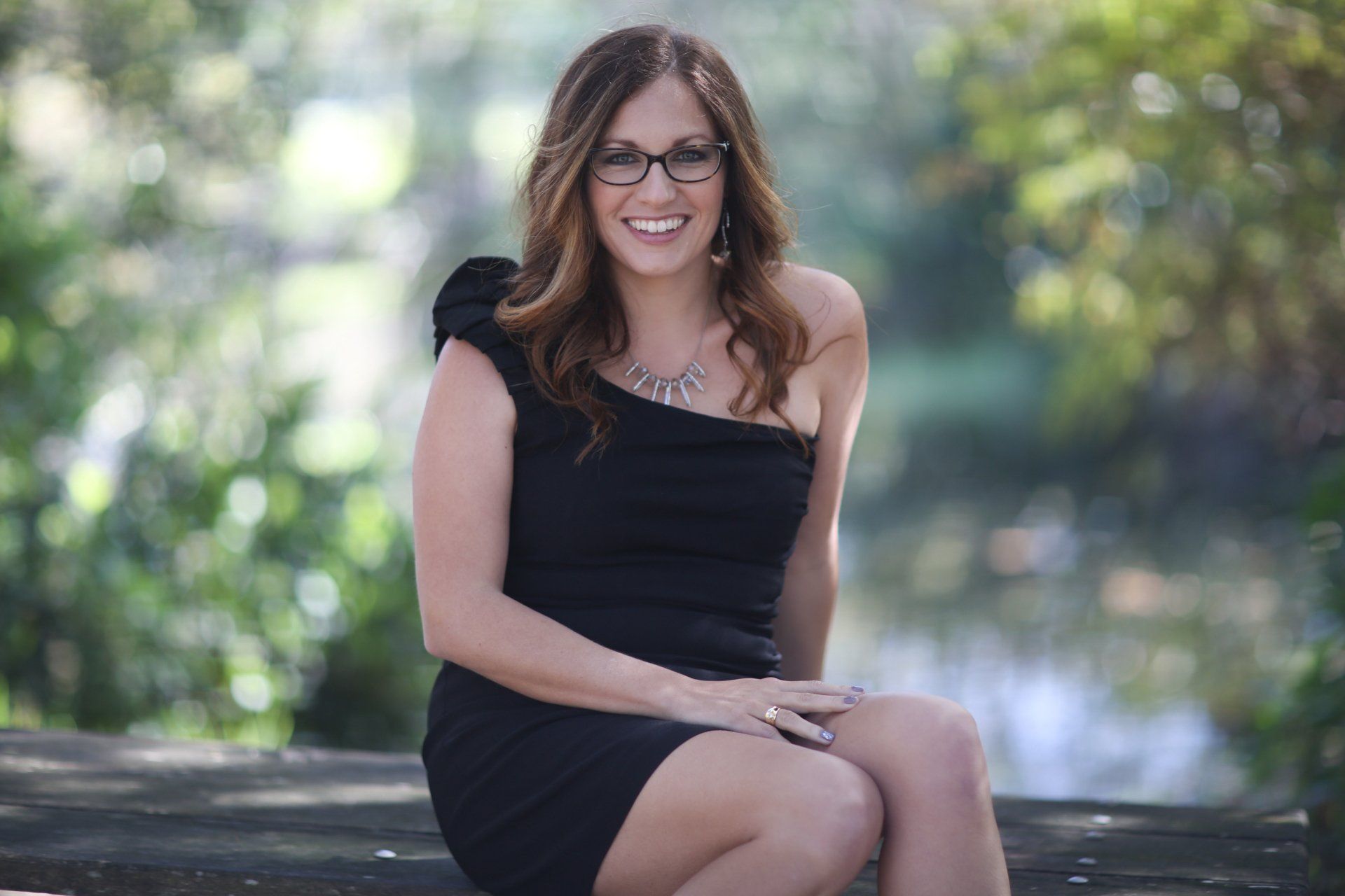 A woman in a black dress and glasses is sitting on a bench.