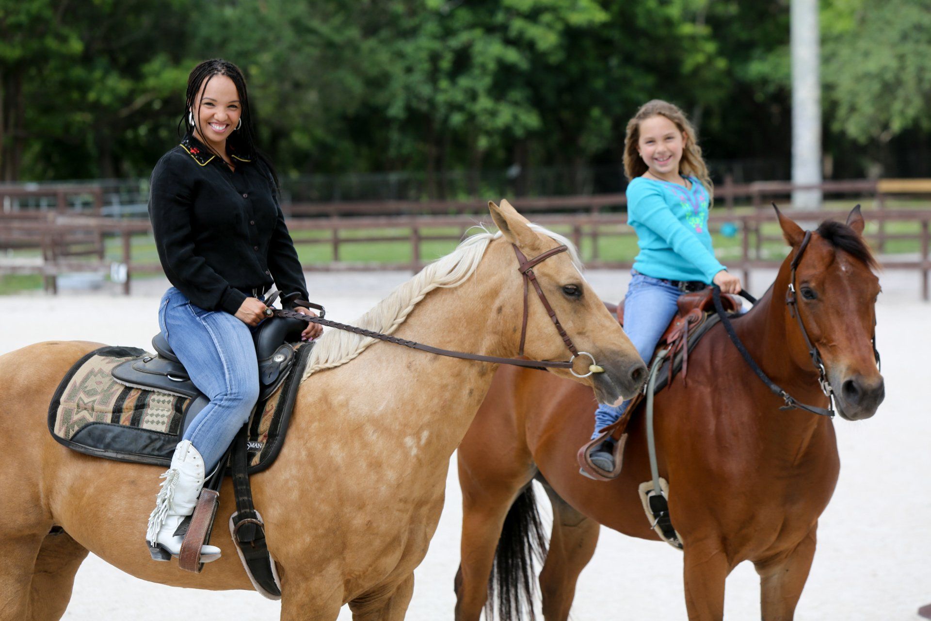 A woman and a little girl are riding horses in a fenced in area.