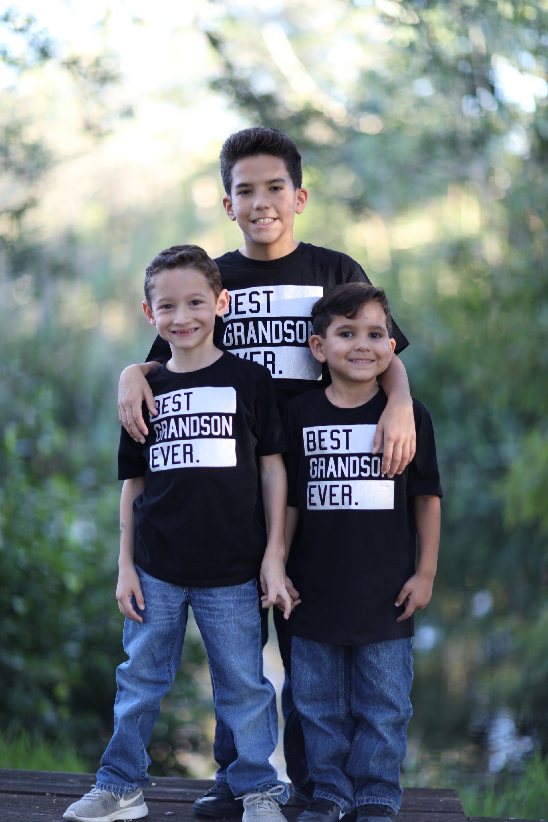 Three young boys wearing black shirts that say `` best grandson ever '' are standing next to each other.