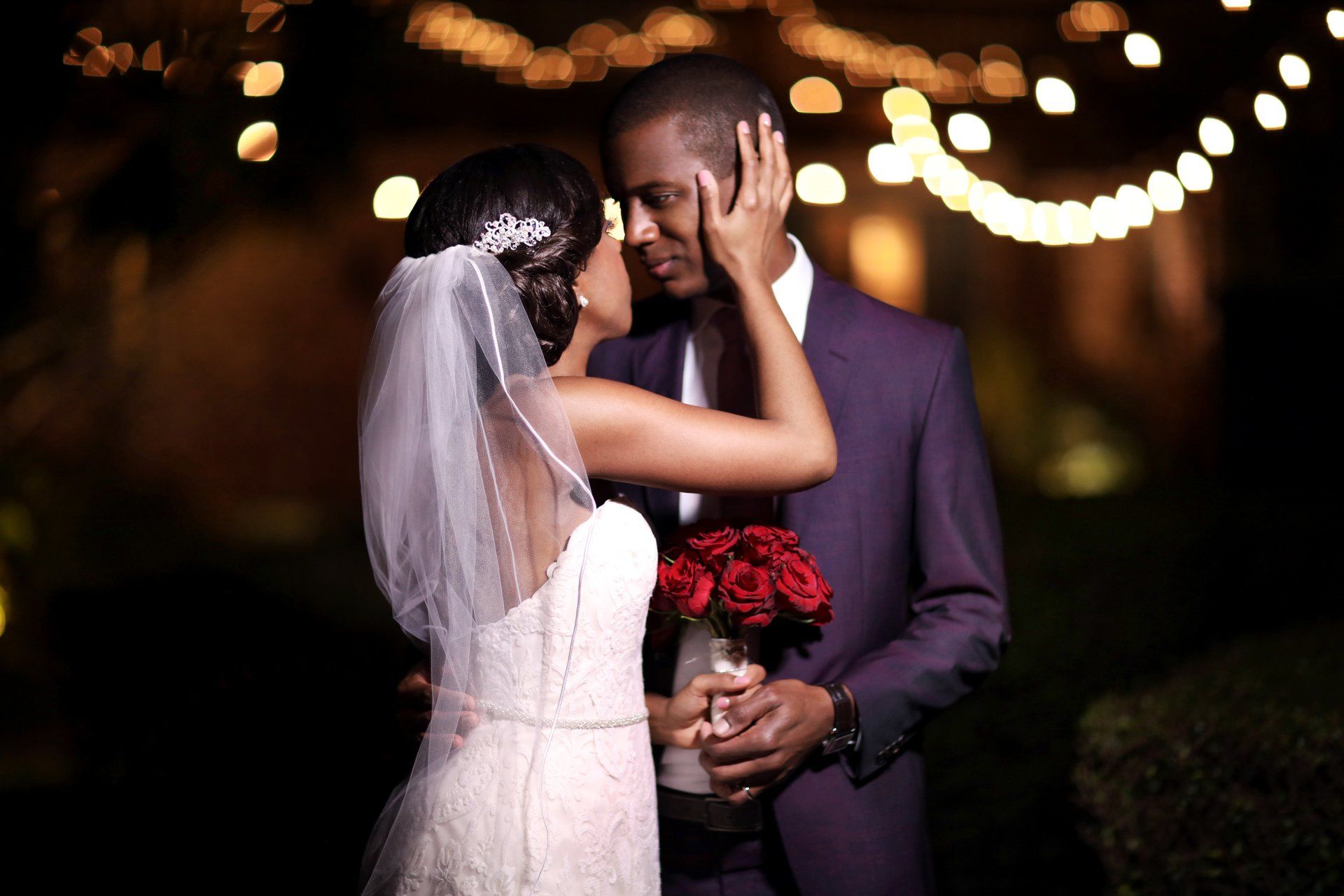 A bride and groom are kissing in front of a string of lights.