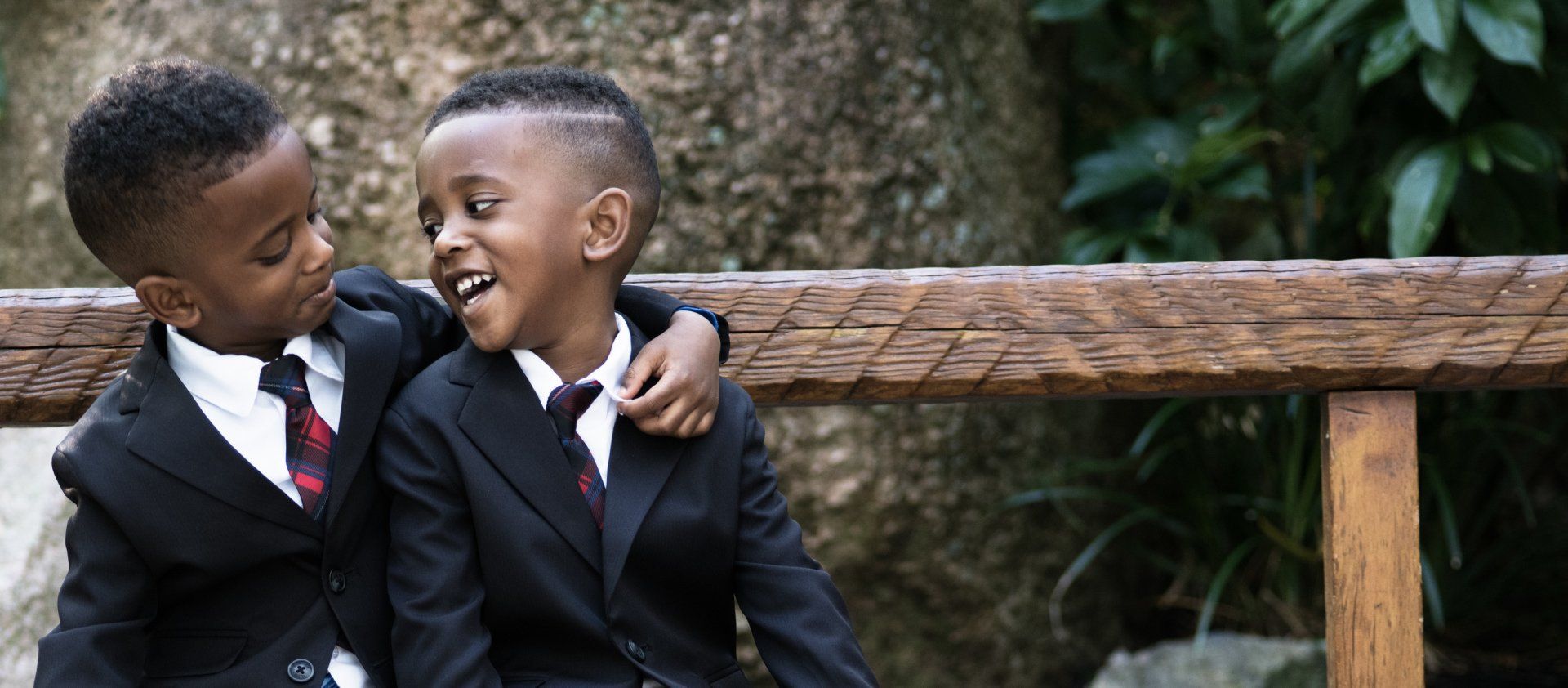 Two young boys in suits and ties are sitting on a wooden bench.