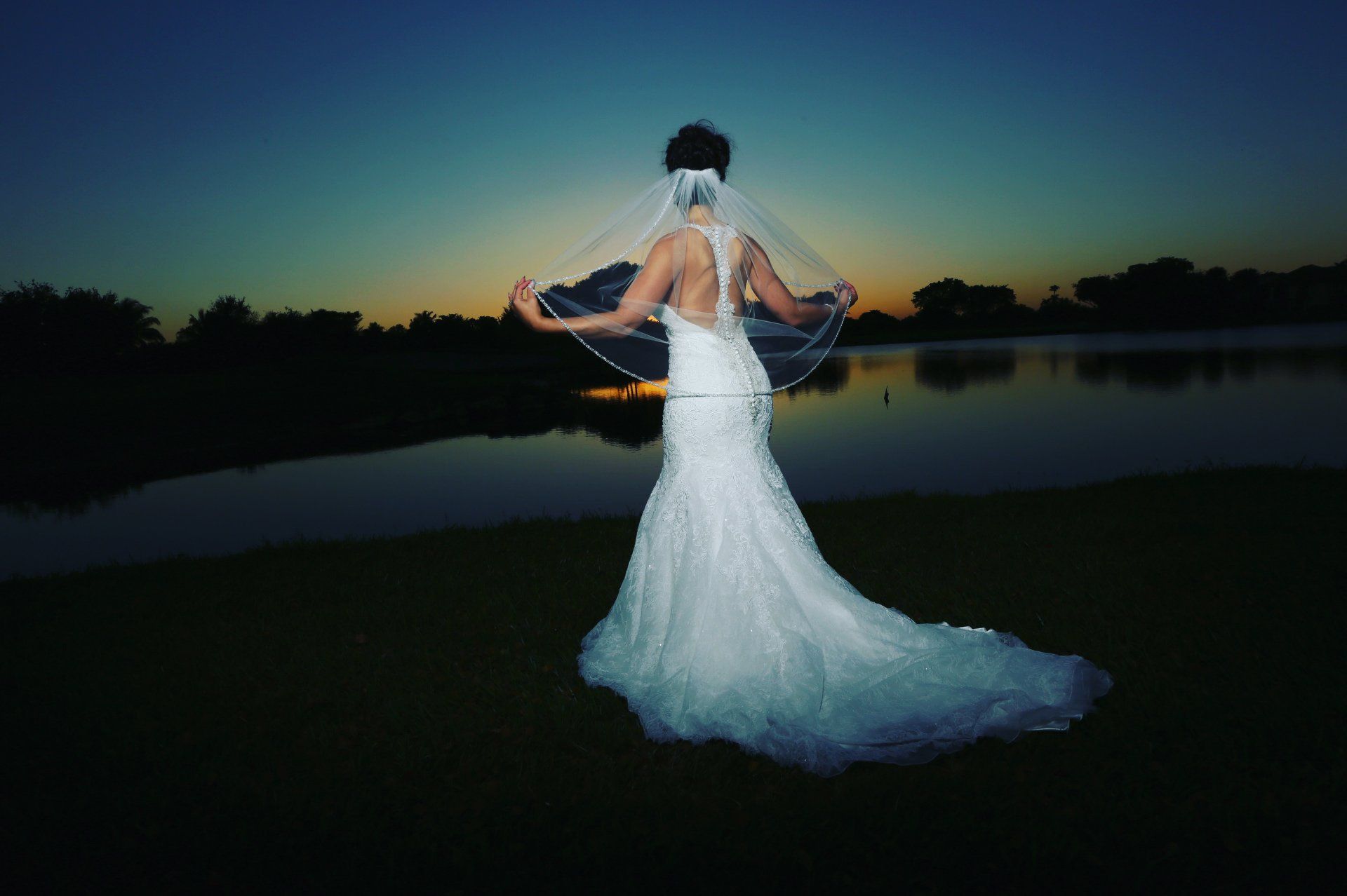 A bride in a wedding dress and veil is standing in front of a lake at sunset.