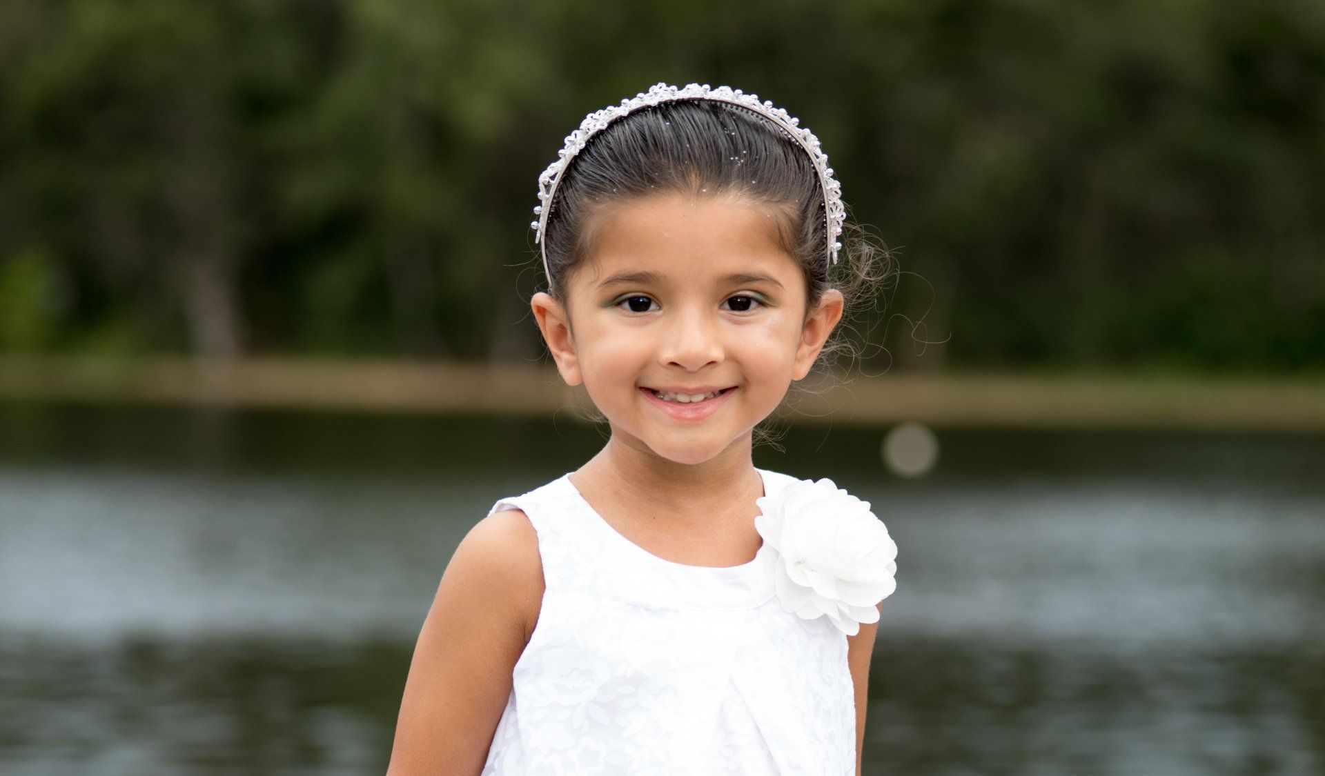 A little girl in a white dress is standing in front of a lake.