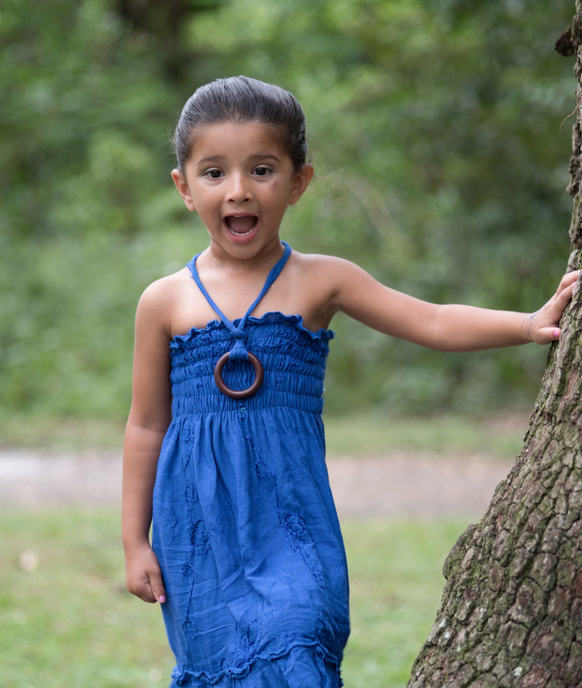 A little girl in a blue dress is leaning against a tree