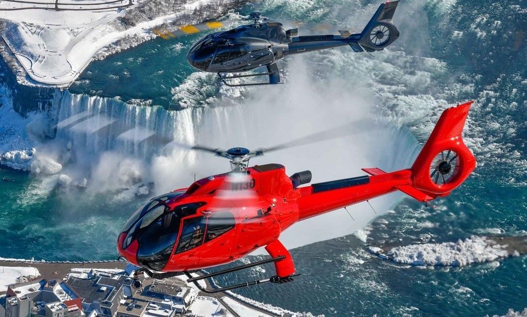 Two helicopters, red and black, flying over Niagara Falls.
