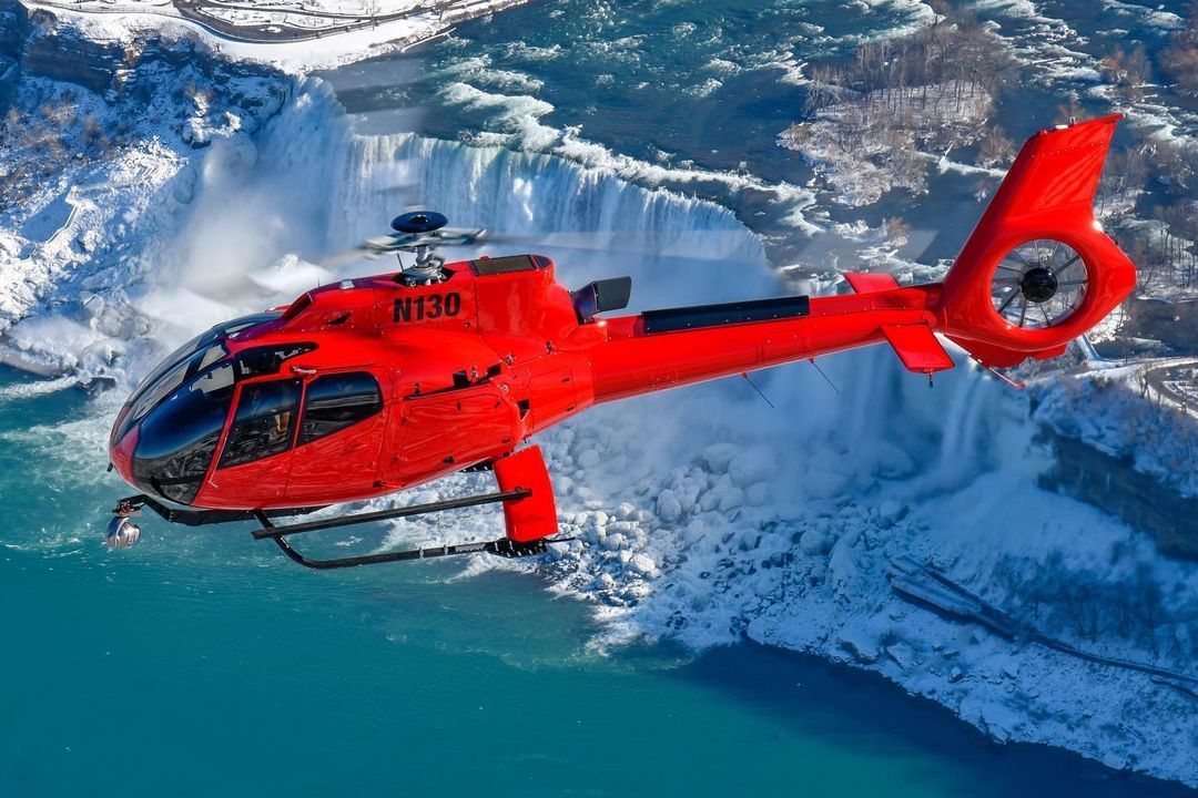 Red helicopter flying over Niagara Falls, with snowy cliffs and turquoise water.