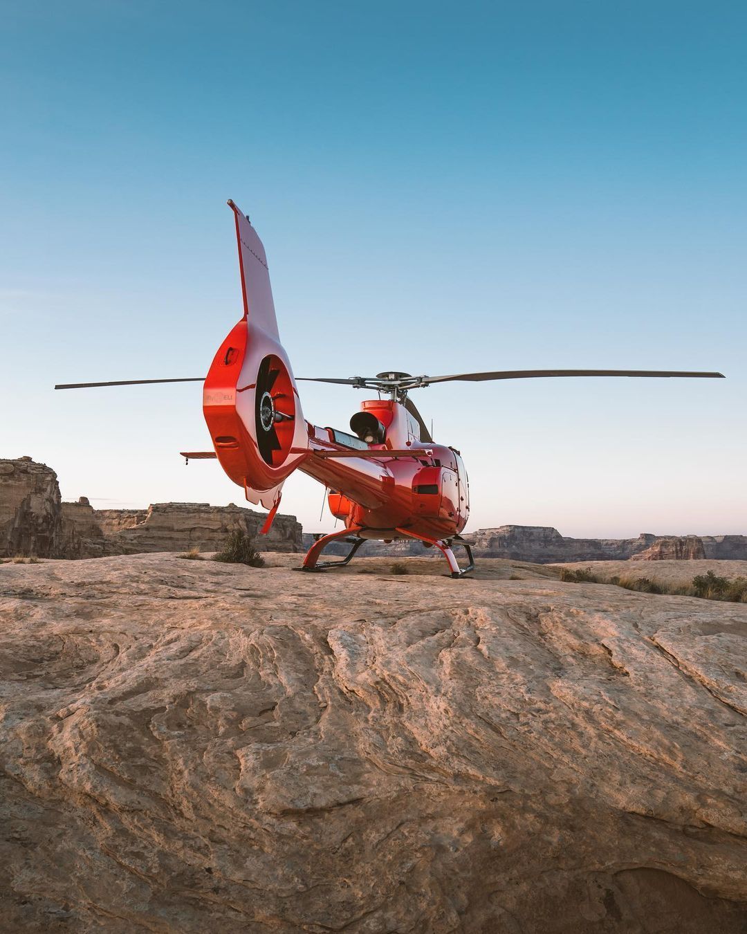 Red helicopter on a rocky, desert-like landscape under a clear blue sky.