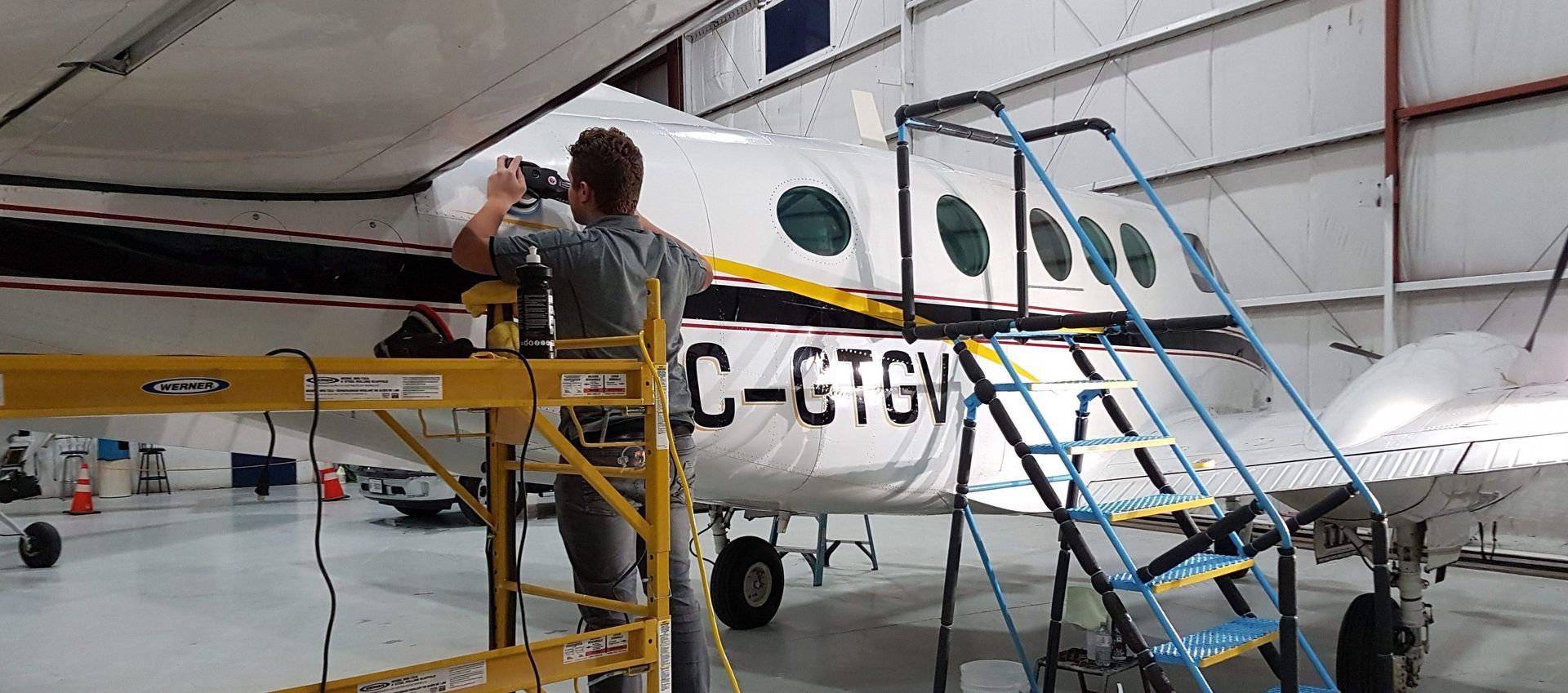 Mechanic working on the side of a white airplane in a hangar, using a yellow lift.