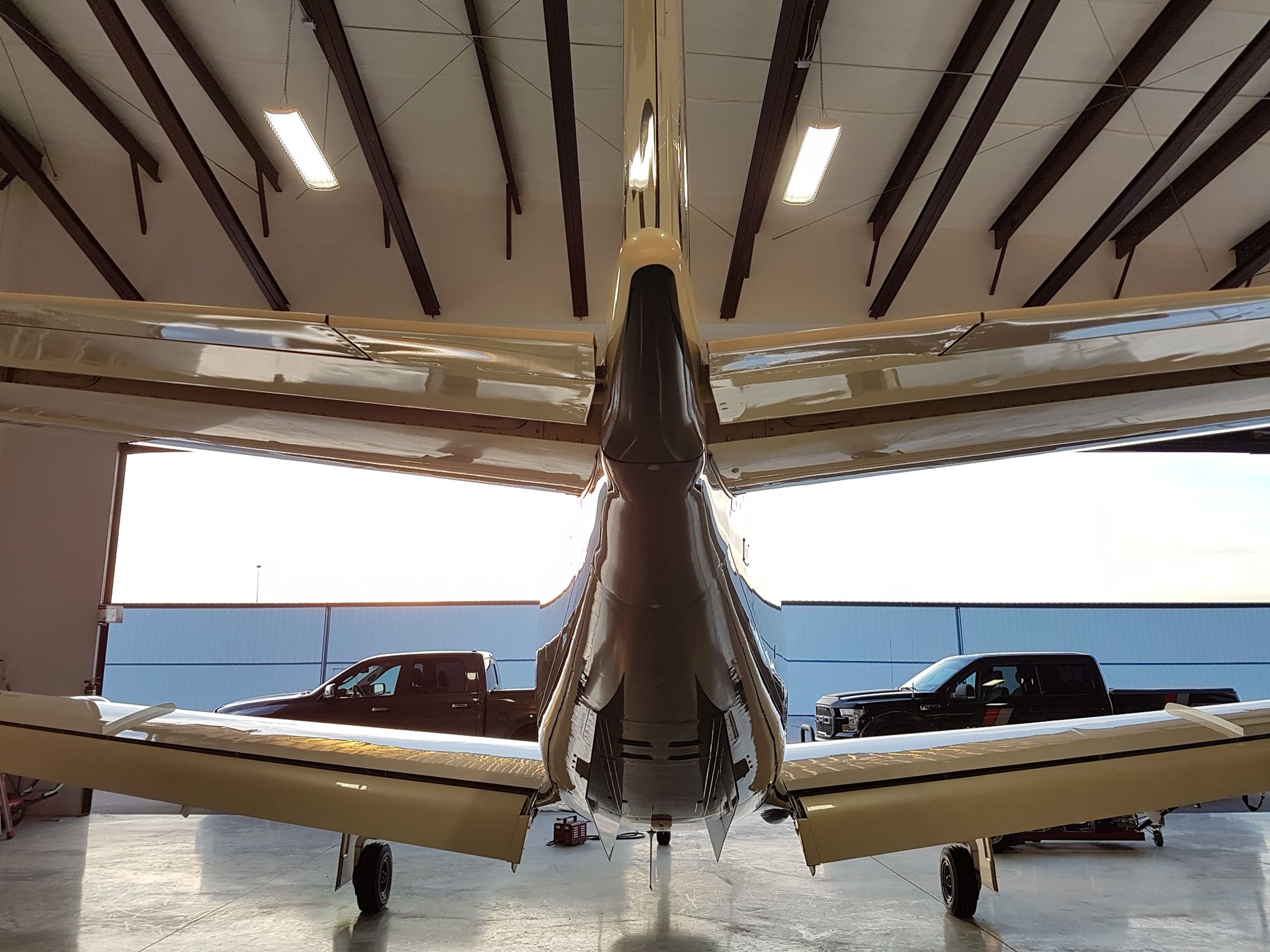 Rear view of a light airplane in a hangar, wings spread. Two trucks are seen outside.