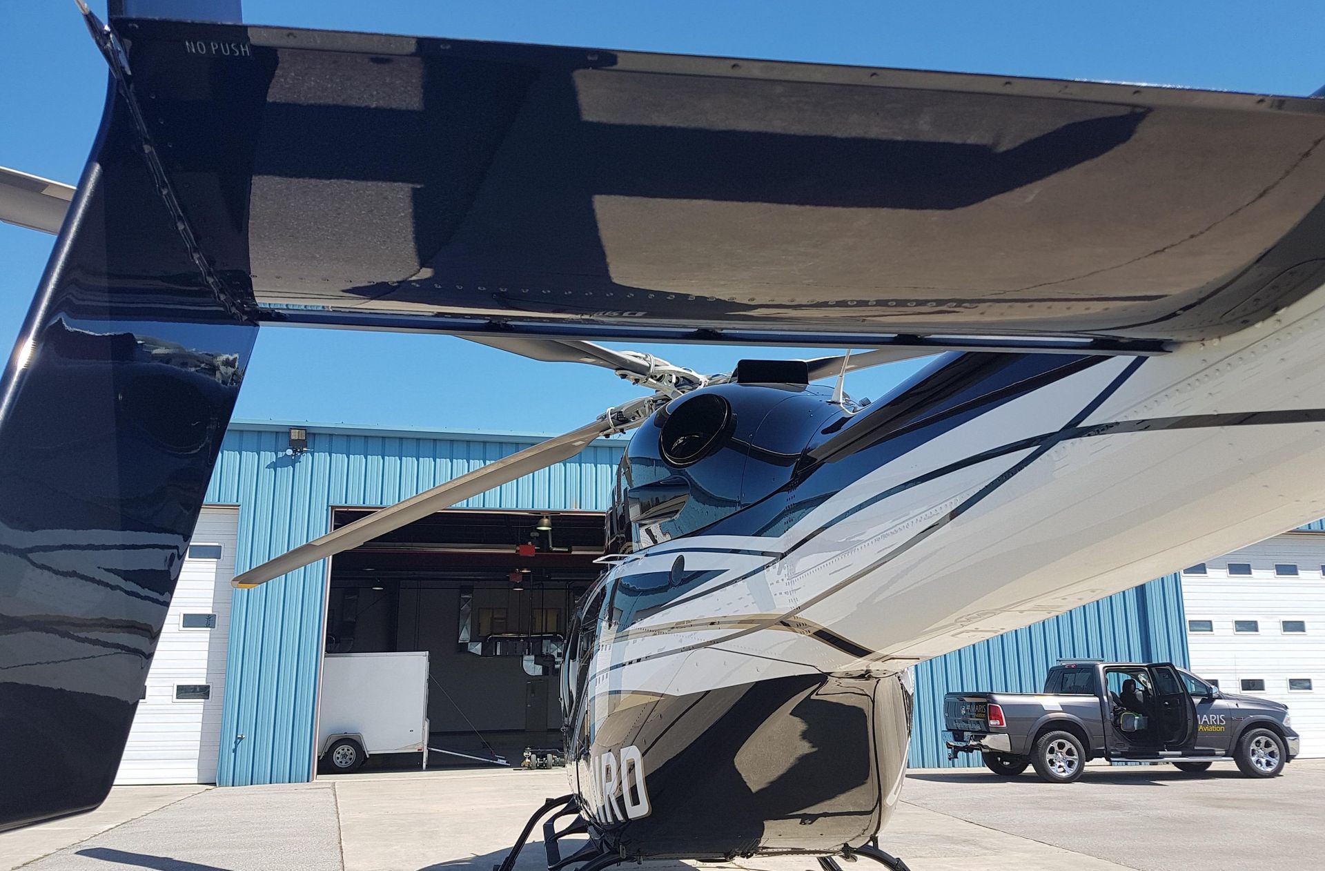 Aircraft tail with propeller; blue and white color scheme; parked near a hangar and a truck.