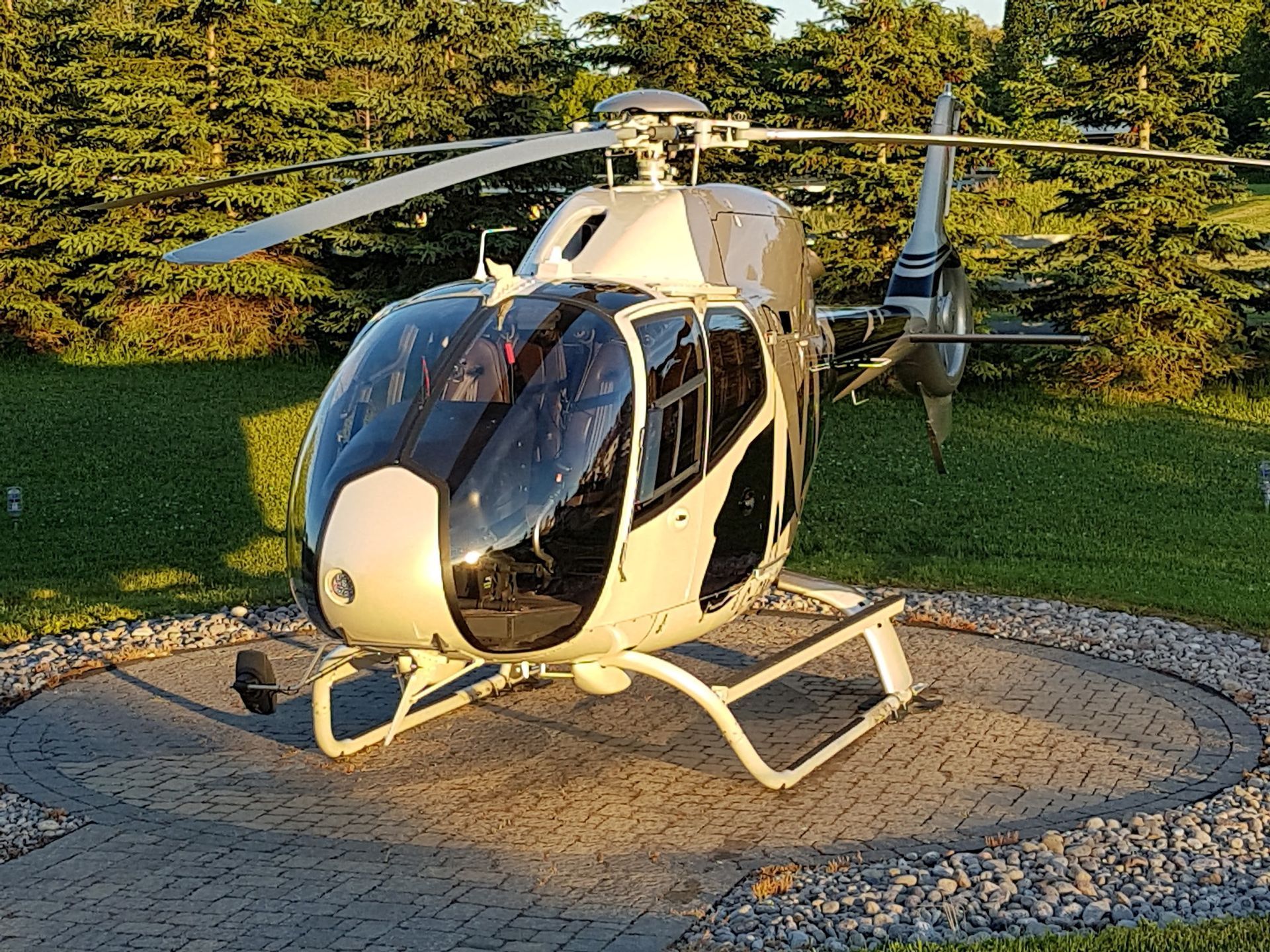 Silver helicopter parked on a circular pad, surrounded by green grass and trees.