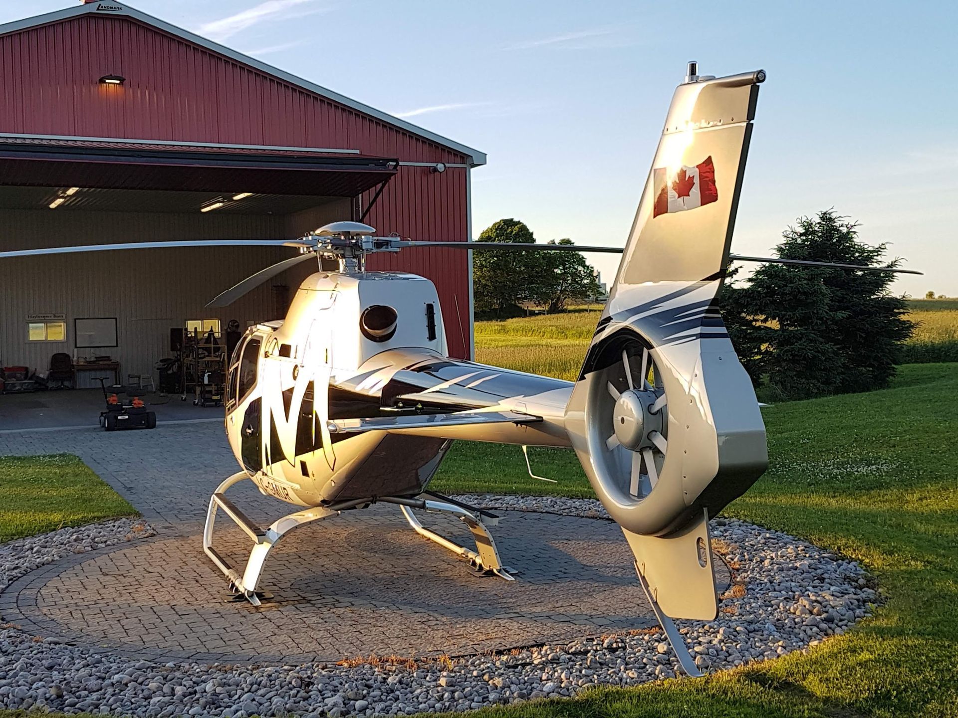 Helicopter on a circular pad in front of a red barn, Canadian flag on the tail.