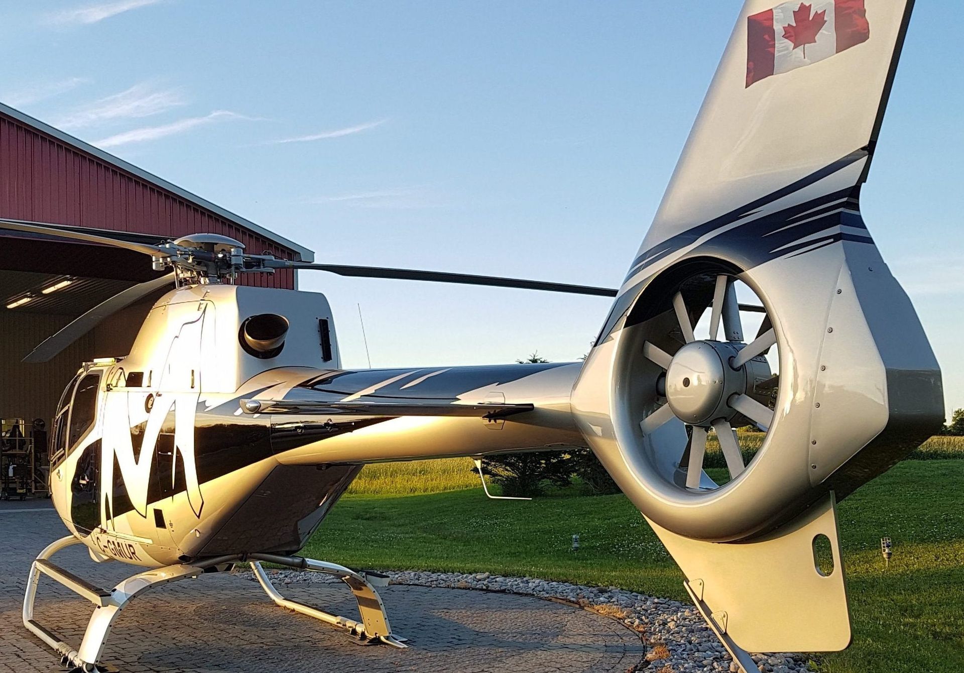 Helicopter with a silver body, Canadian flag, and tail rotor, parked on a paved area with a red building in the background.