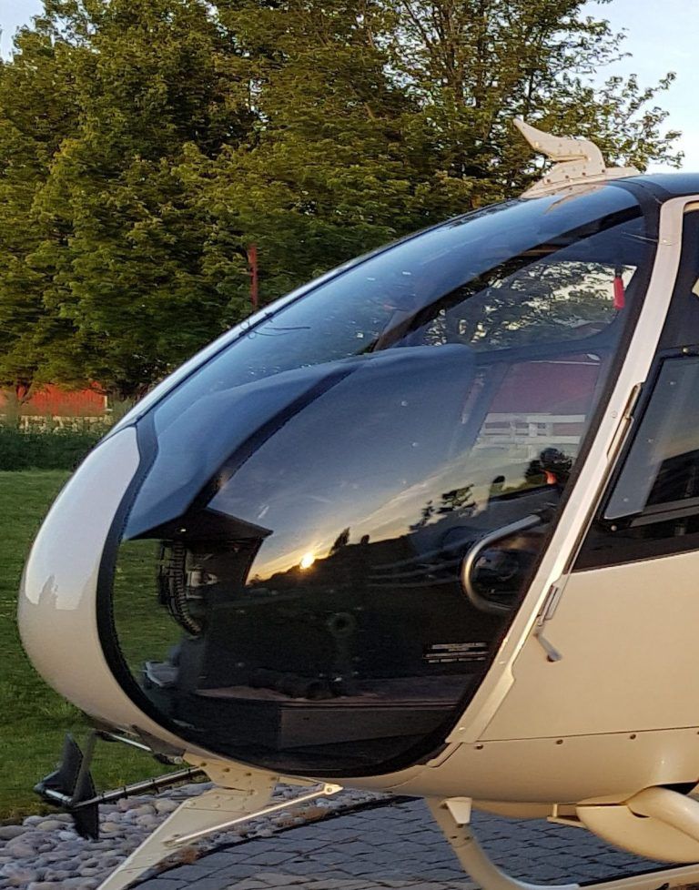 Helicopter cockpit, clear windshield reflecting a sunset. White fuselage, set against trees and a grassy area.