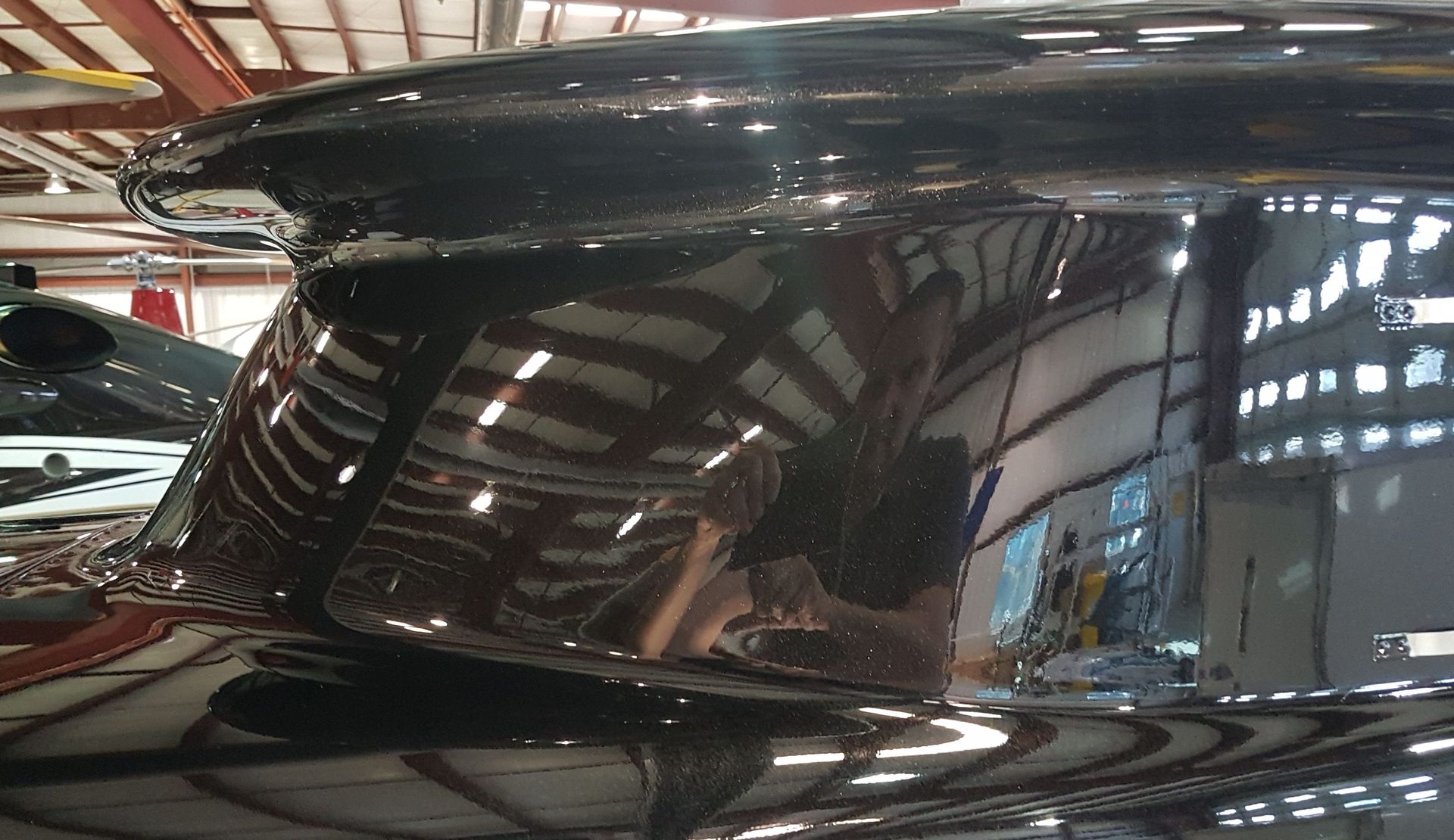 Close-up of a glossy, dark aircraft surface reflecting a hangar interior with lights and other planes.