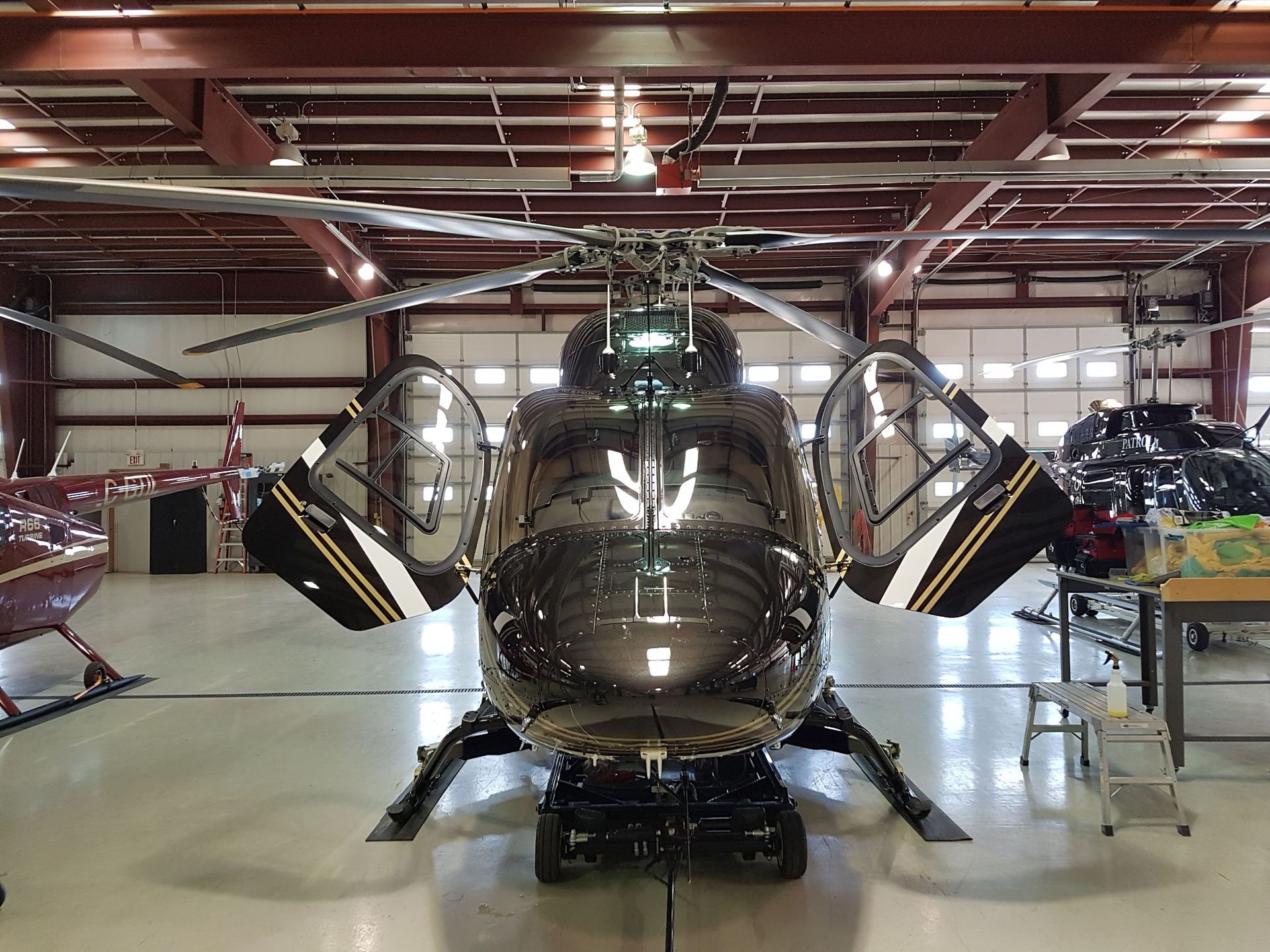 Black helicopter in hangar, doors open, front view. Brown and white trim, other aircraft in background.