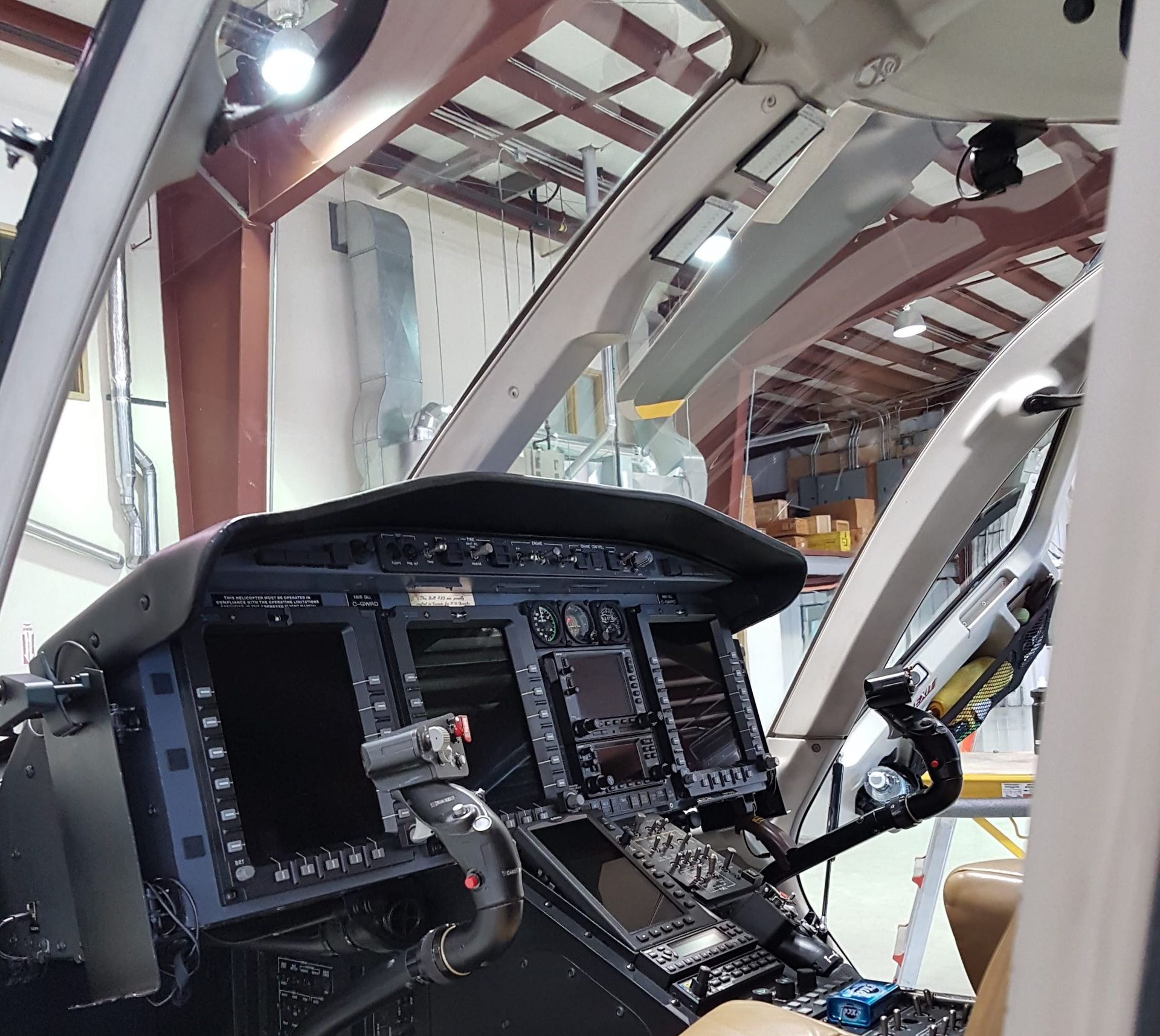 Helicopter cockpit interior: dark instrument panel, screens, and controls, seen from a passenger's seat.
