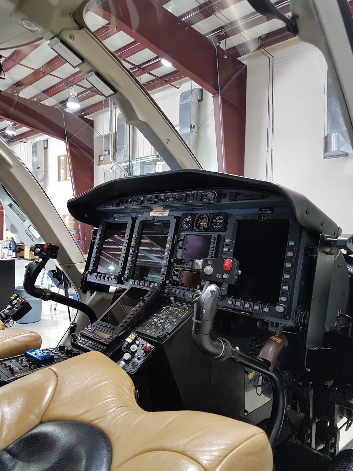 Helicopter cockpit interior: digital displays, control panel, beige seats. In a hangar.