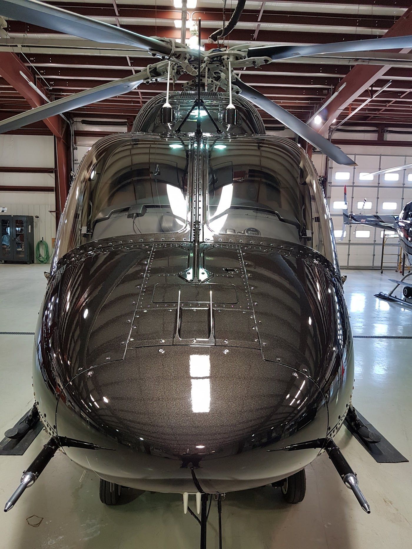 Black helicopter in a hangar, reflective surface. Rotor blades, landing gear, and a garage door visible.
