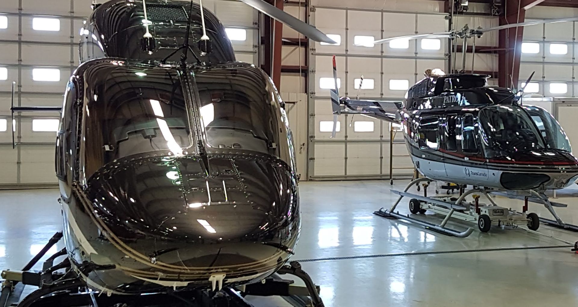 Two black helicopters inside a hangar with a reflective floor and large windows.