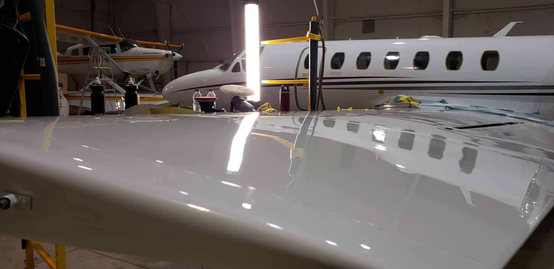 A white airplane wing with shiny reflection, in a hangar with another airplane.