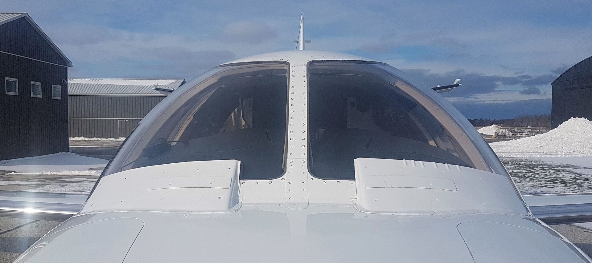 A white airplane cockpit with a clear dome-shaped windshield, and another airplane in the background. Snowy environment.