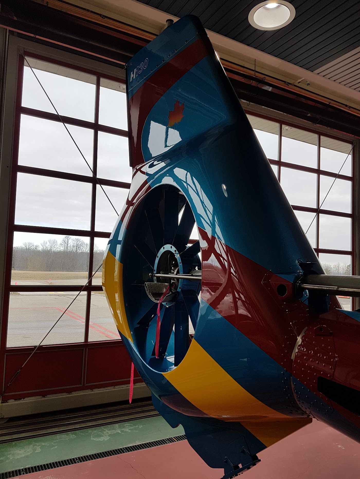 Blue, red, and yellow tail rotor of an aircraft. It is inside a hangar with a window and a cloudy sky in the background.