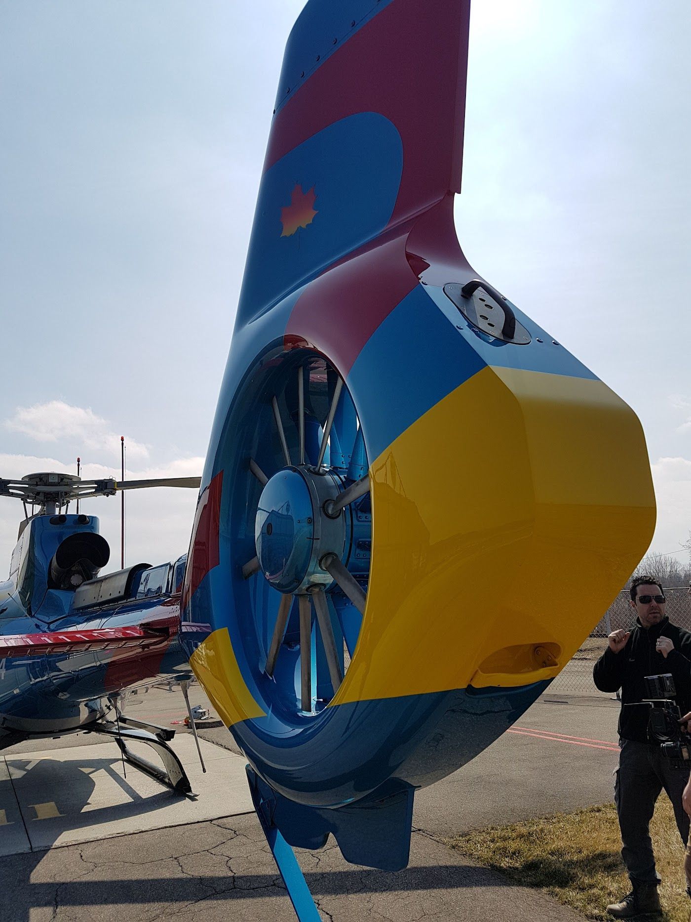 Tail of a blue, yellow, and red helicopter with a person in uniform standing nearby.