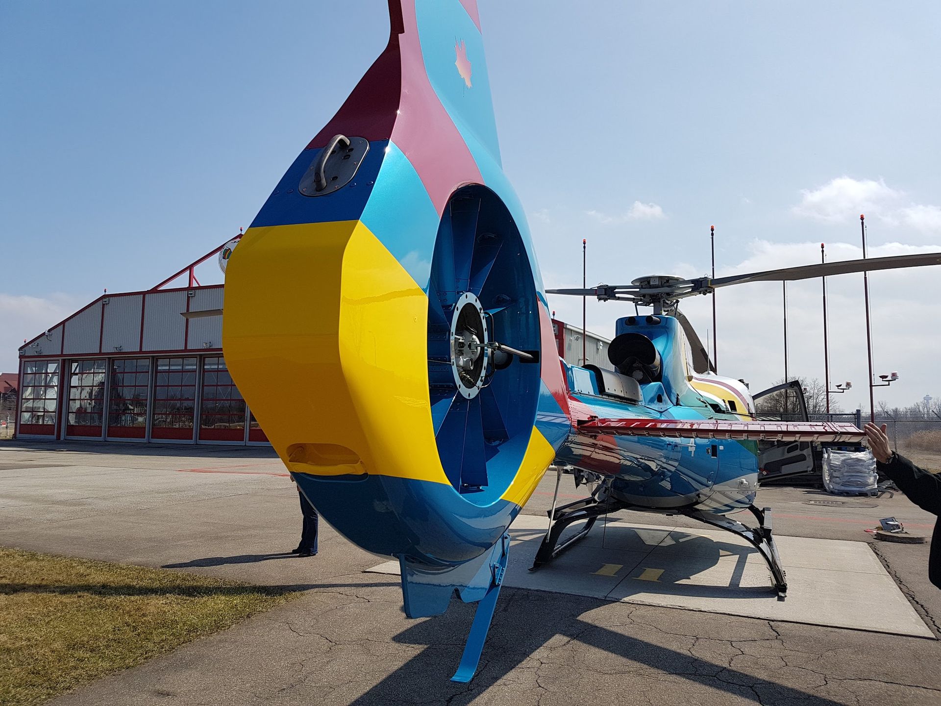 Rear view of a blue, yellow, and red helicopter on a tarmac, near a building on a sunny day.