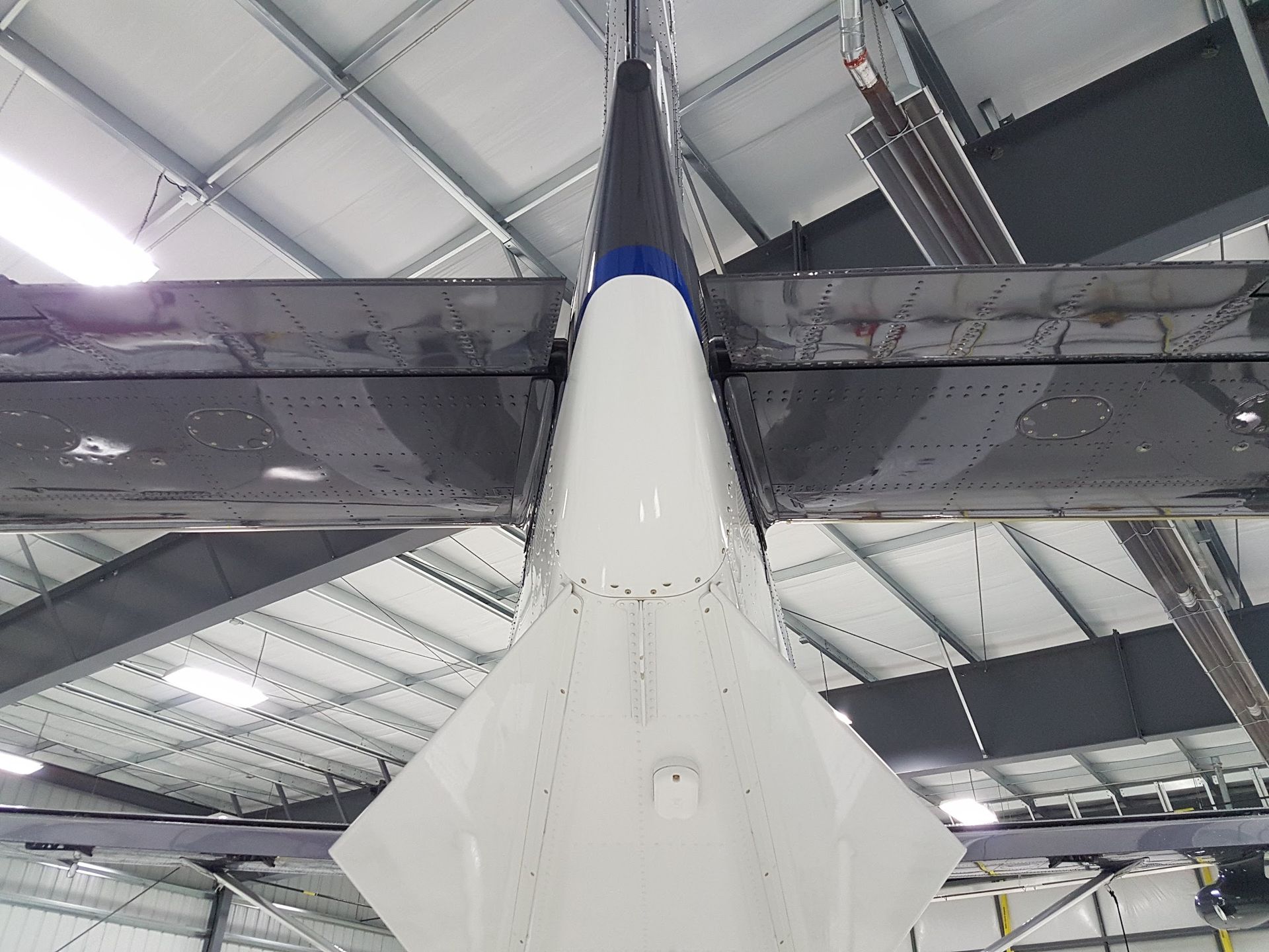 Tail of a white and black airplane inside a hangar. Dark wings extend from the base of the tail.