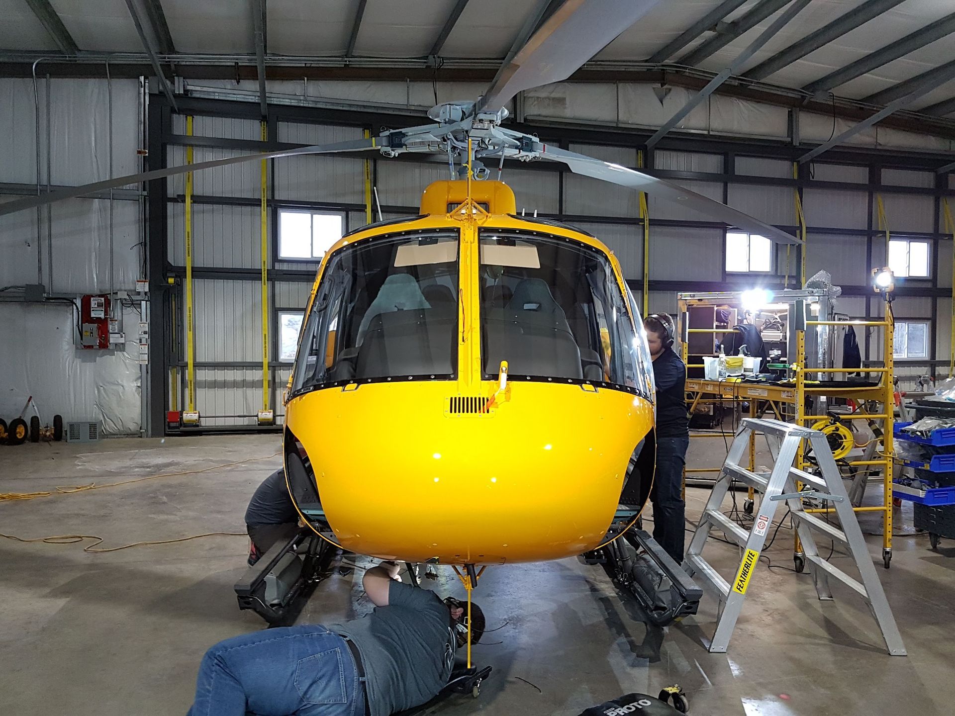 Yellow helicopter being worked on inside a hangar. Two people are working underneath, one on a creeper.