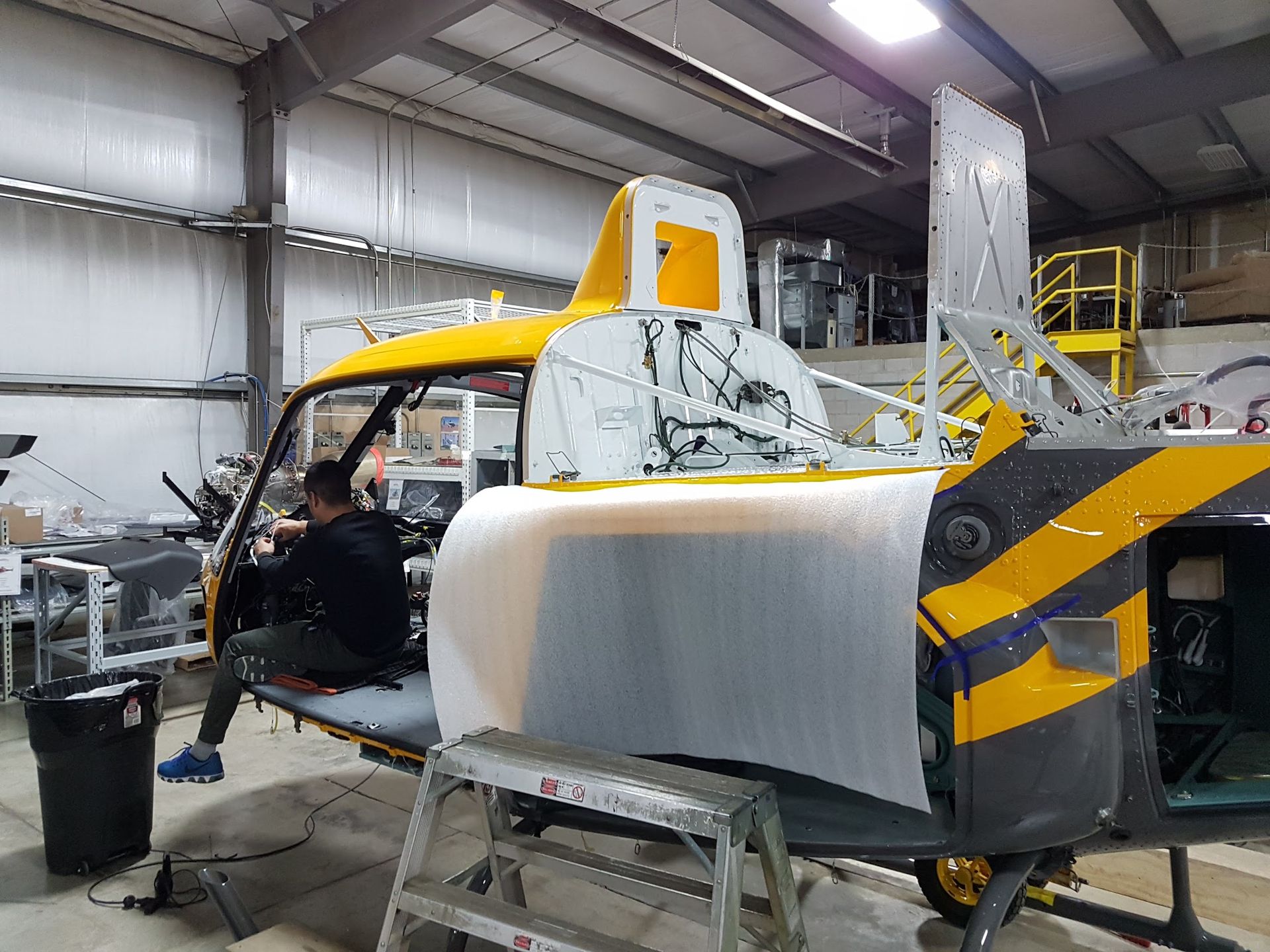 Helicopter undergoing maintenance inside a hangar. A person works on the cockpit. Yellow and black markings are visible.