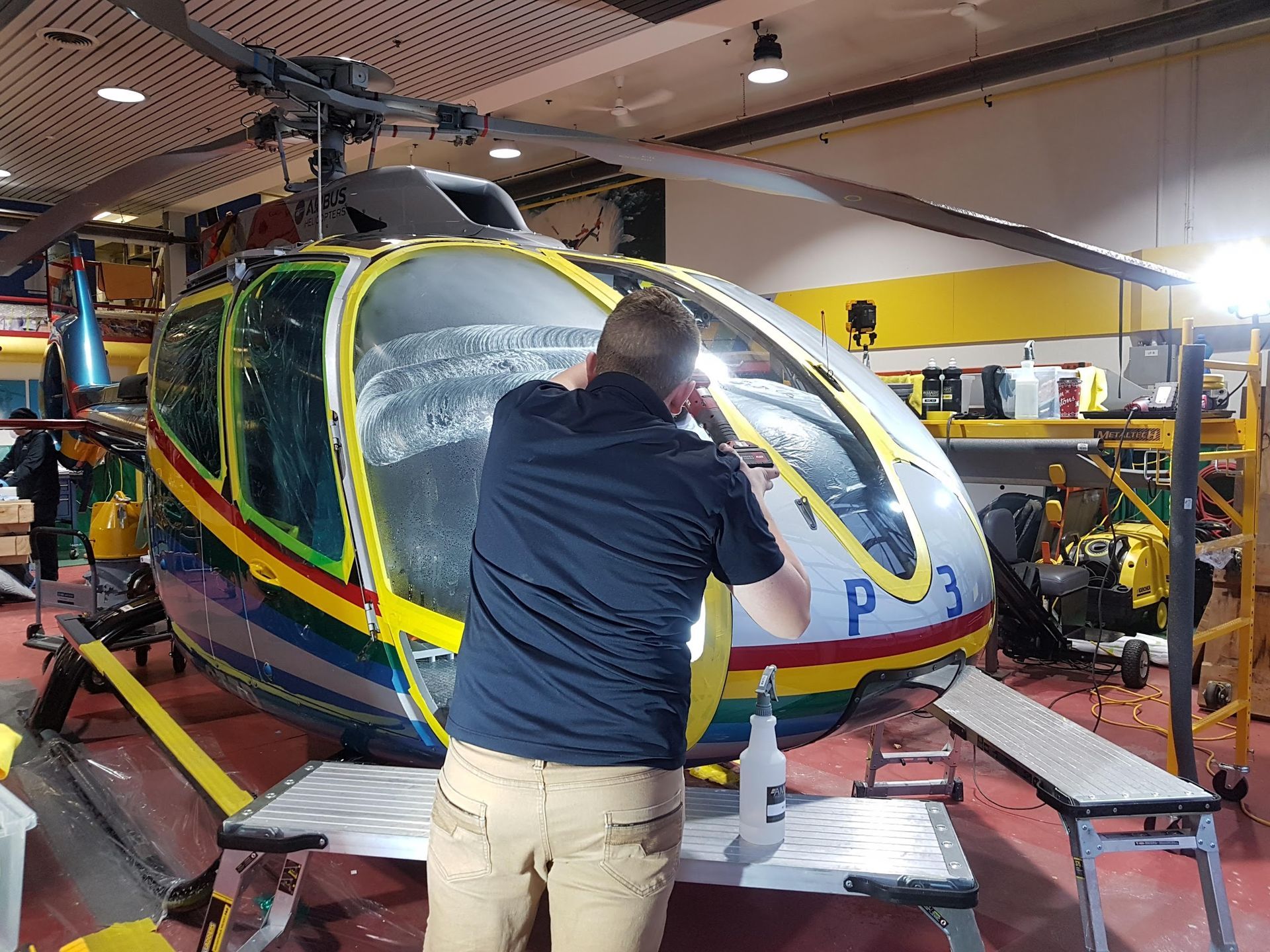 A person works on a helicopter, applying material to the windshield, inside a hangar.