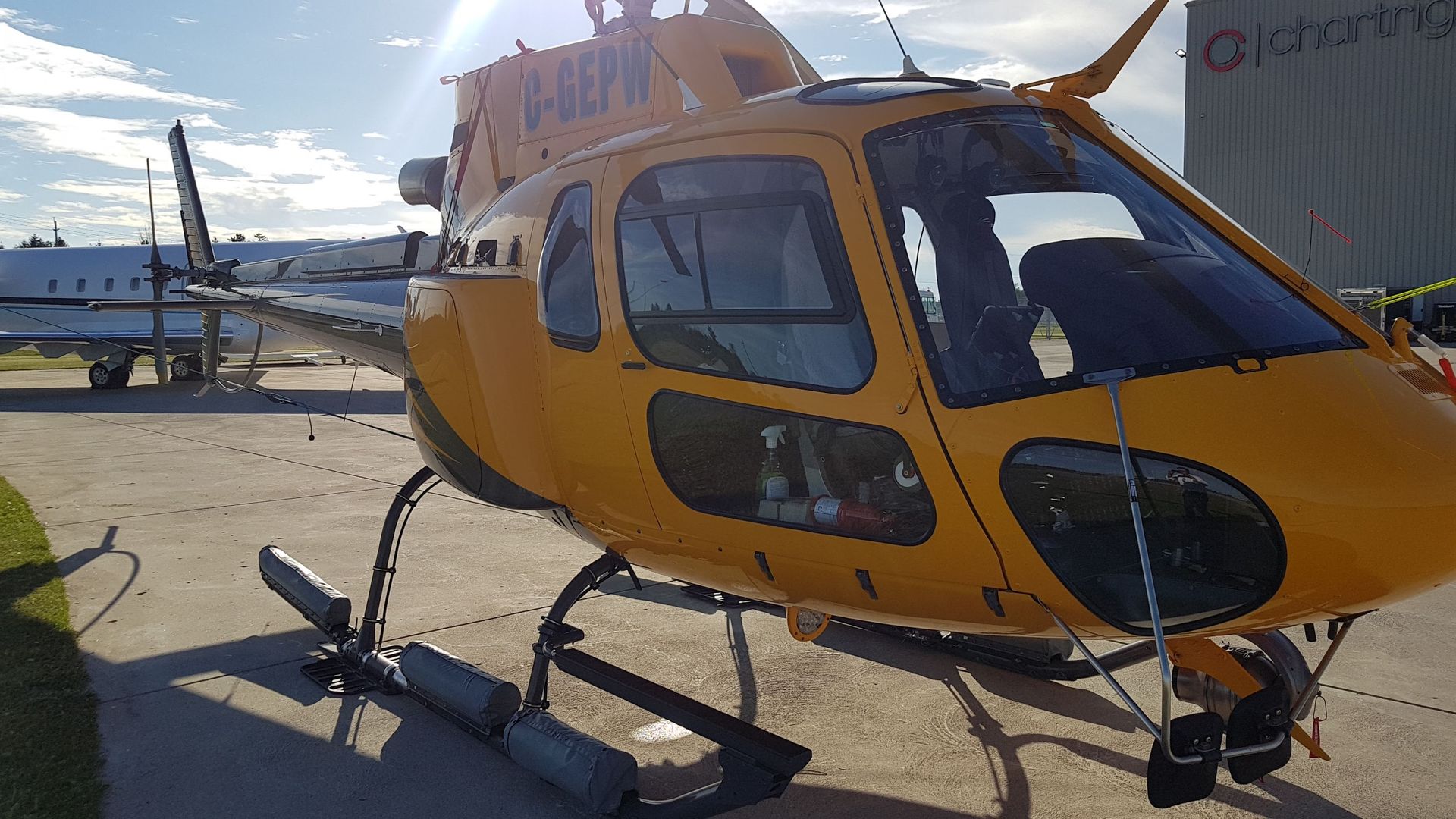 Yellow helicopter on tarmac with another plane and hangar in background.
