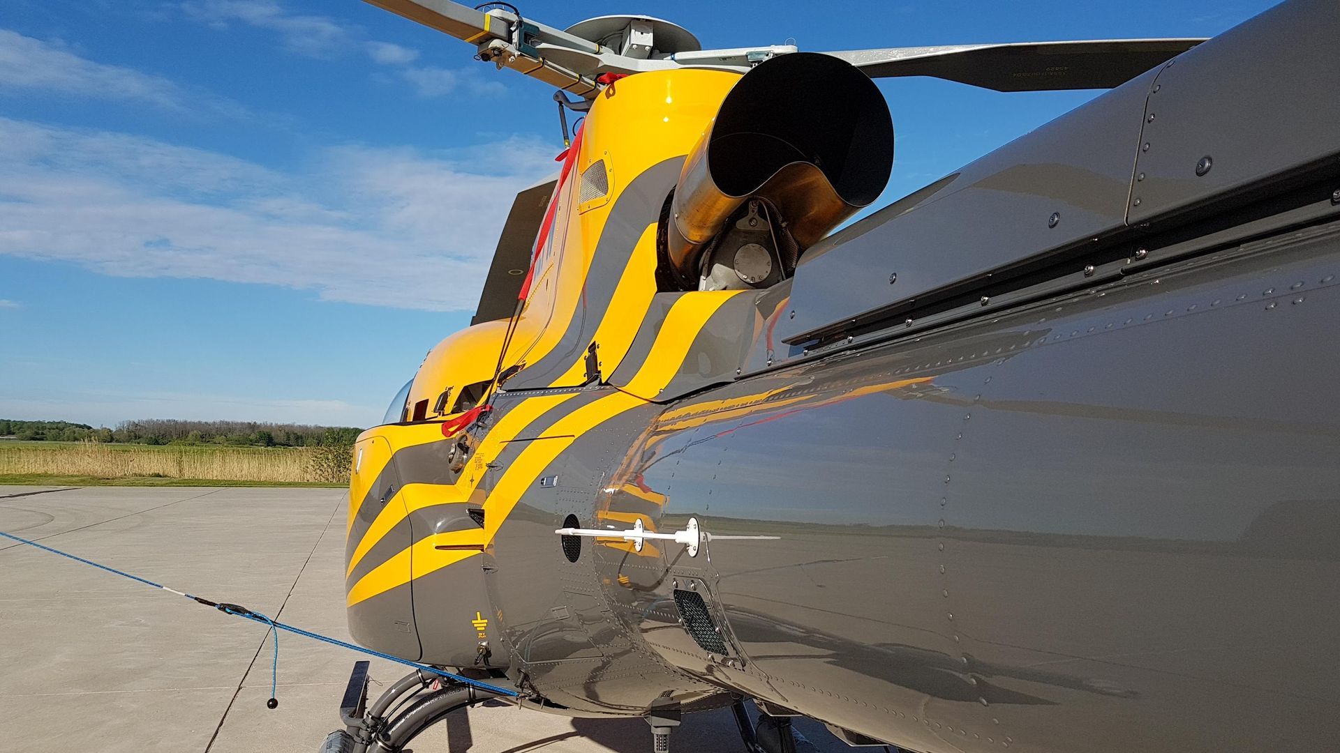 Close-up of a gray helicopter's tail, with yellow and black tiger-stripe accents on a sunny day.