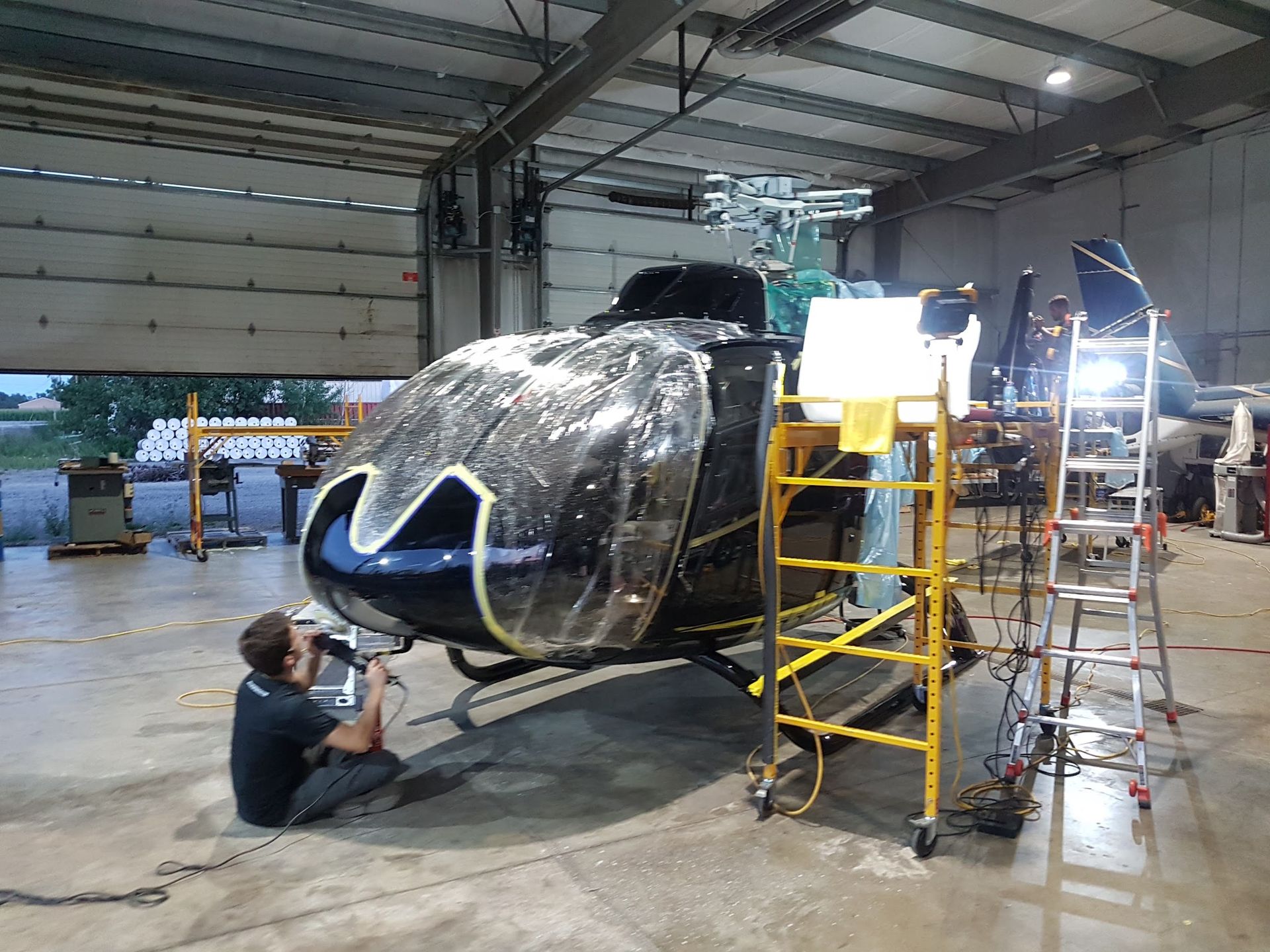 Person working on a helicopter inside a hangar. The helicopter is black and blue. There's scaffolding and a ladder nearby.
