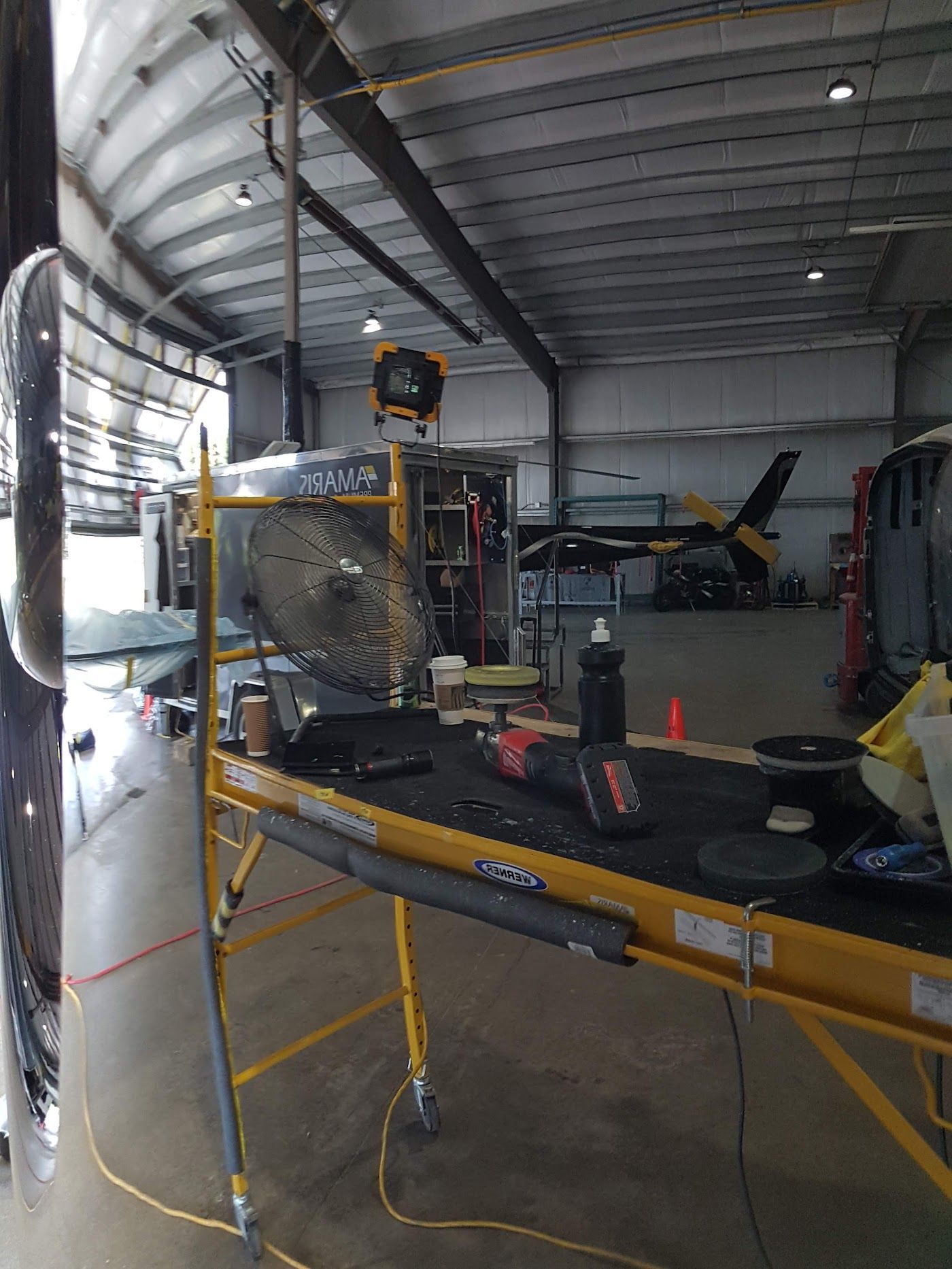 A black vehicle being worked on inside a garage. Scaffolding holds tools and a fan.