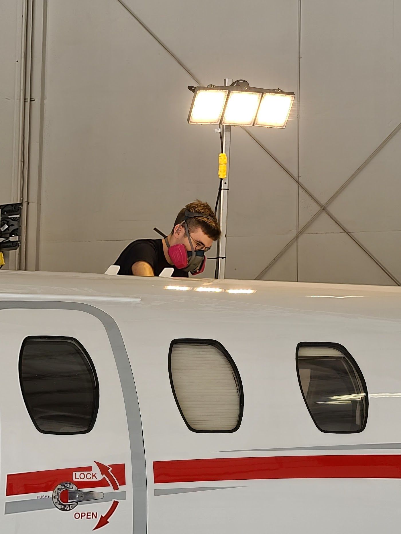 Person wearing mask working on airplane exterior, lit by overhead lights in a hangar.
