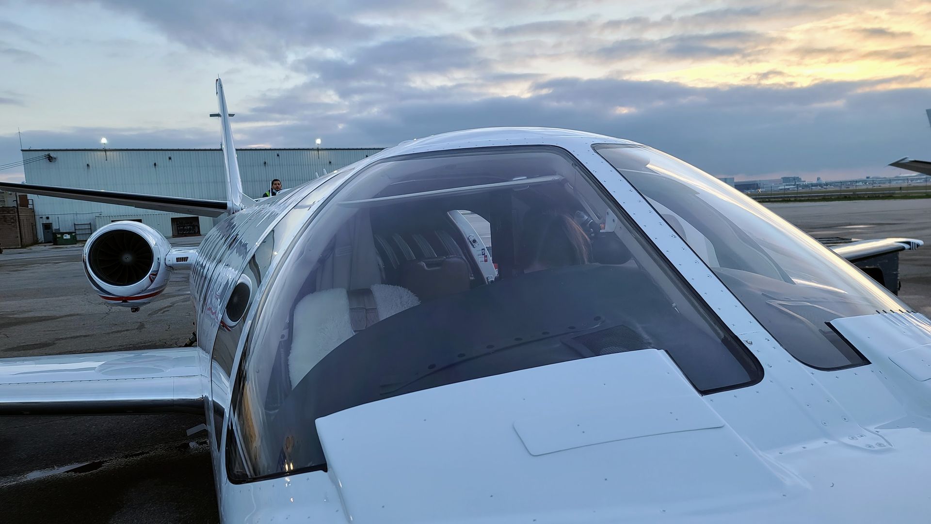 White jet cockpit, parked on tarmac. Pilot visible inside. Cloudy sky in the background.