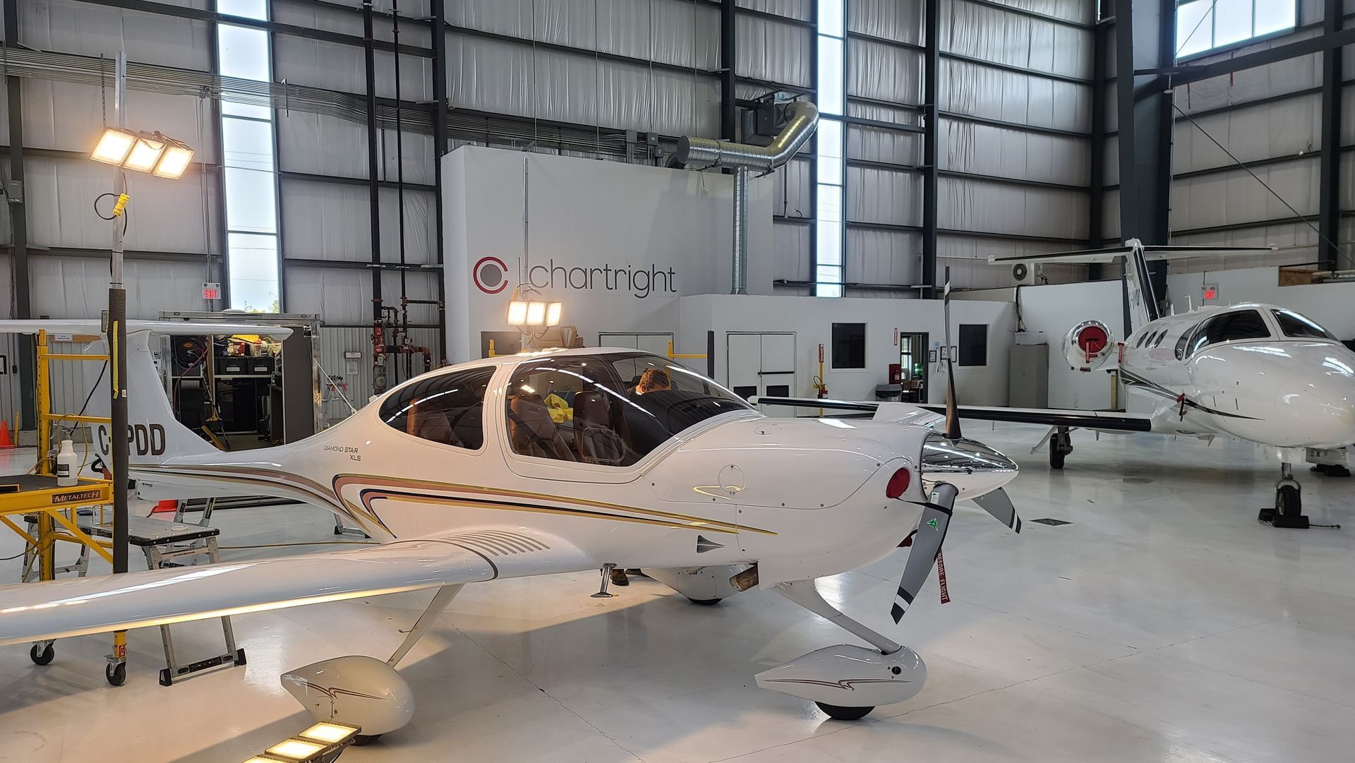 White airplane inside a hangar, another plane visible to the right. Fluorescent lights and a sign for