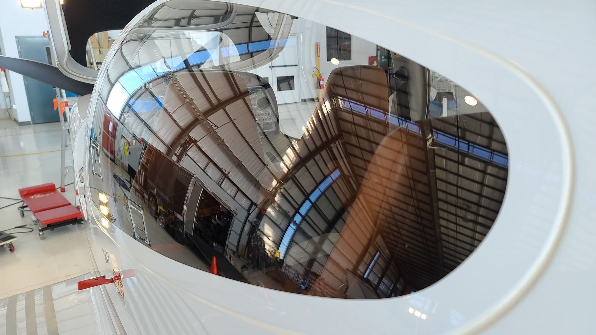 Reflections in a curved cockpit window, showing a hangar interior with red items and metal structures.